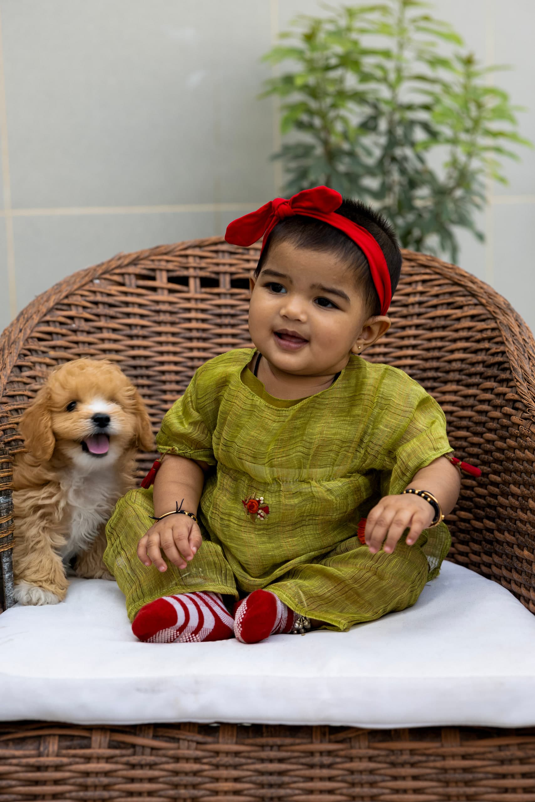 Baby in green looks to the camera, a small cream puppy sitting on the cushion beside her