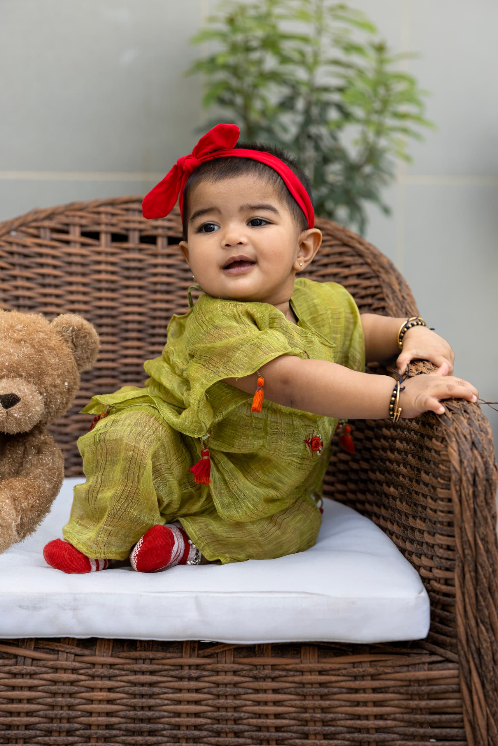 Baby turning to grip the arm of the cane chair, red bow headband, teddy at the edge of frame