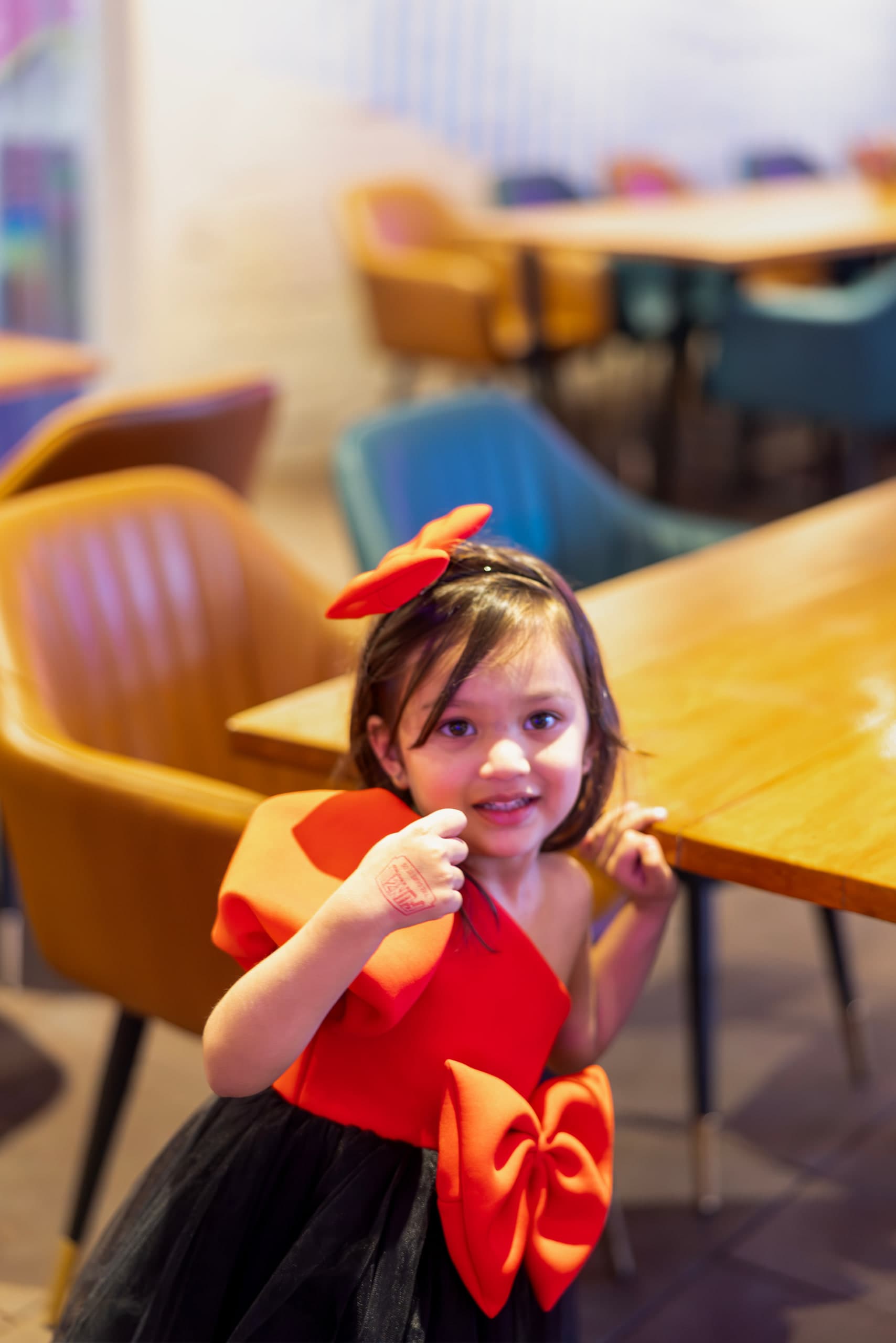 A girl in a red and black party dress mid-step between café tables, hands raised