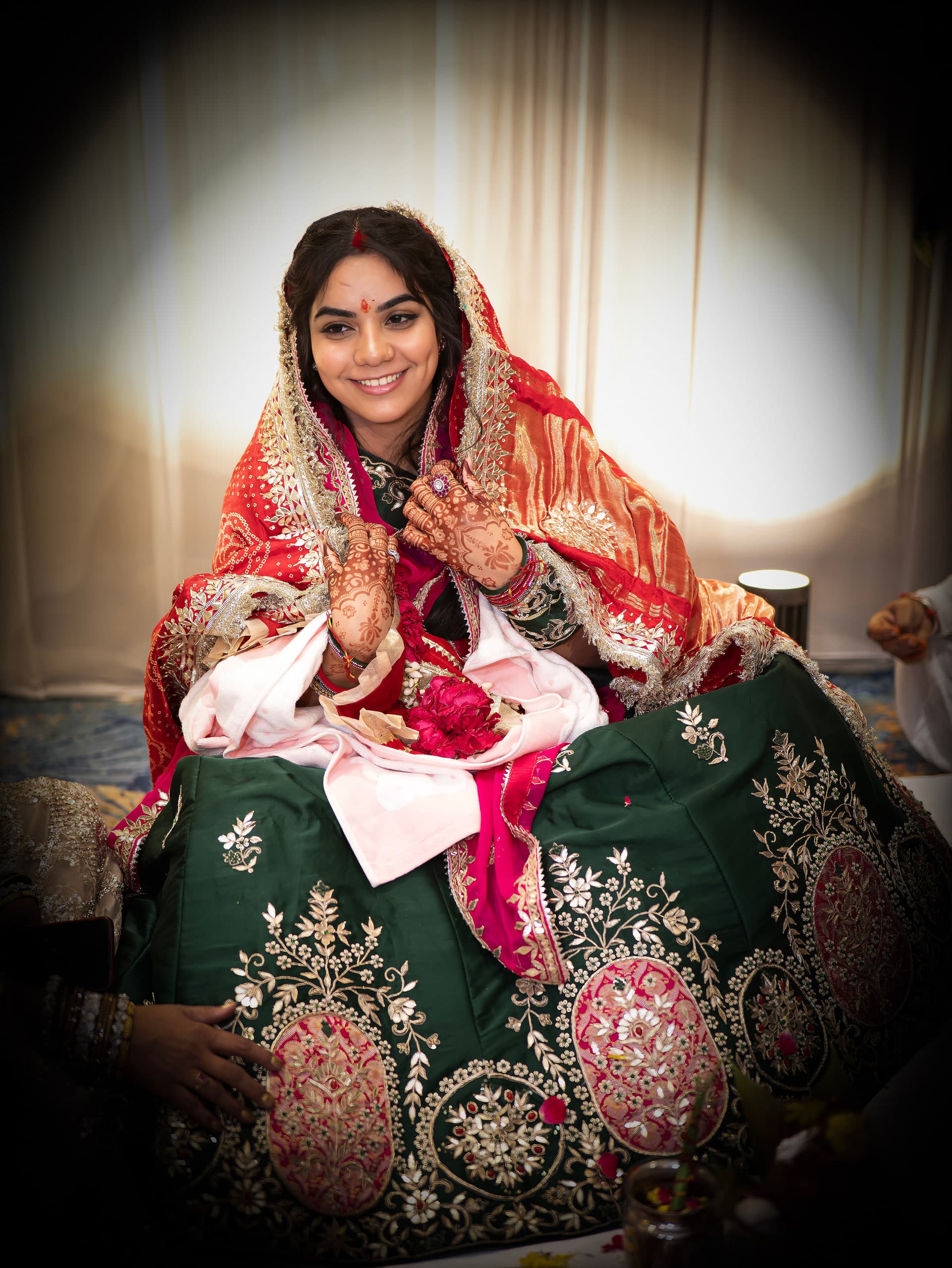 Bride seated in a dark green lehenga and red dupatta, hand raised, warm light on her