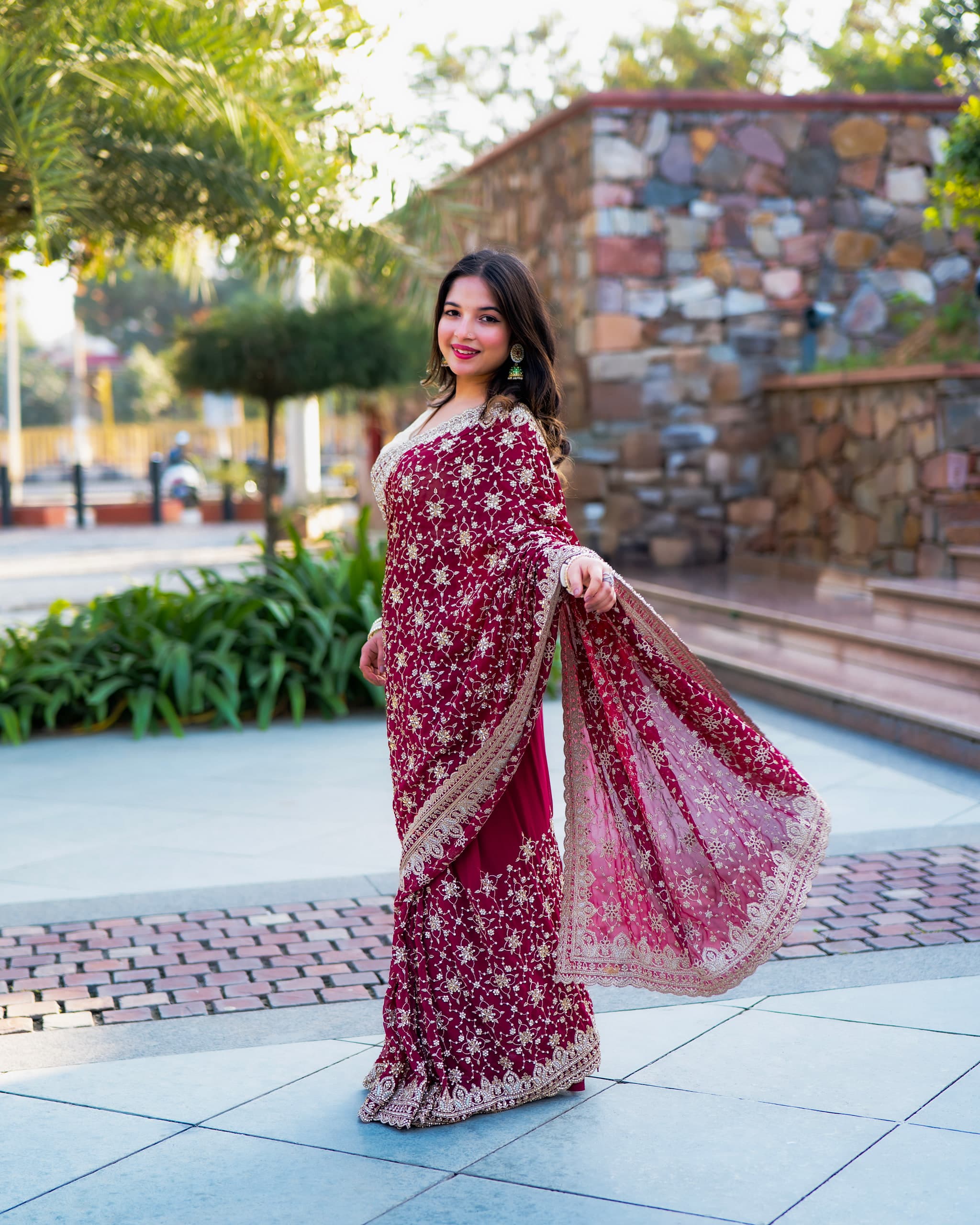 Model mid-turn on a paved courtyard in a red and gold embroidered saree, stone wall and planting behind
