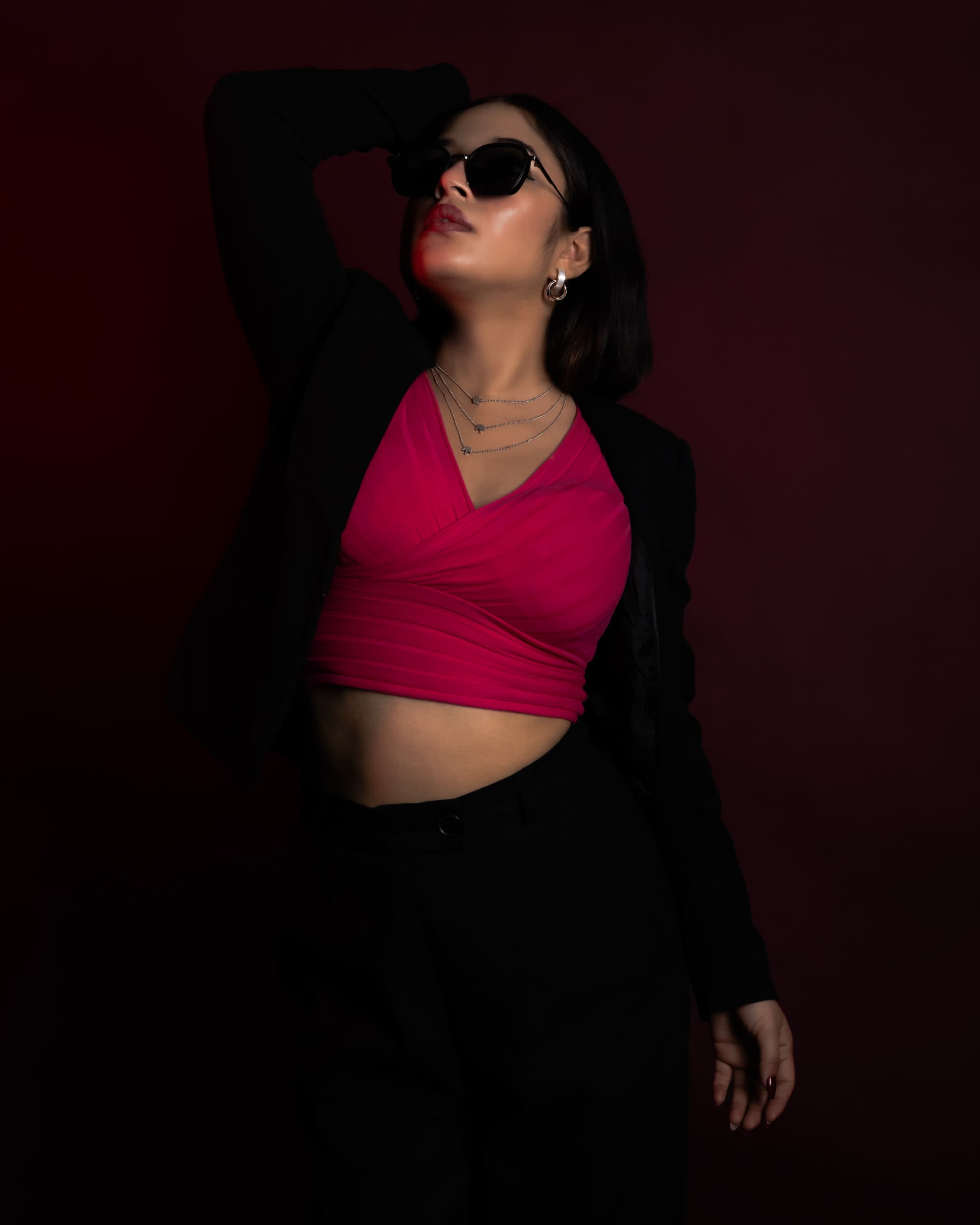 Full-length on crimson, one arm raised behind the head, pink cropped top under an open black blazer