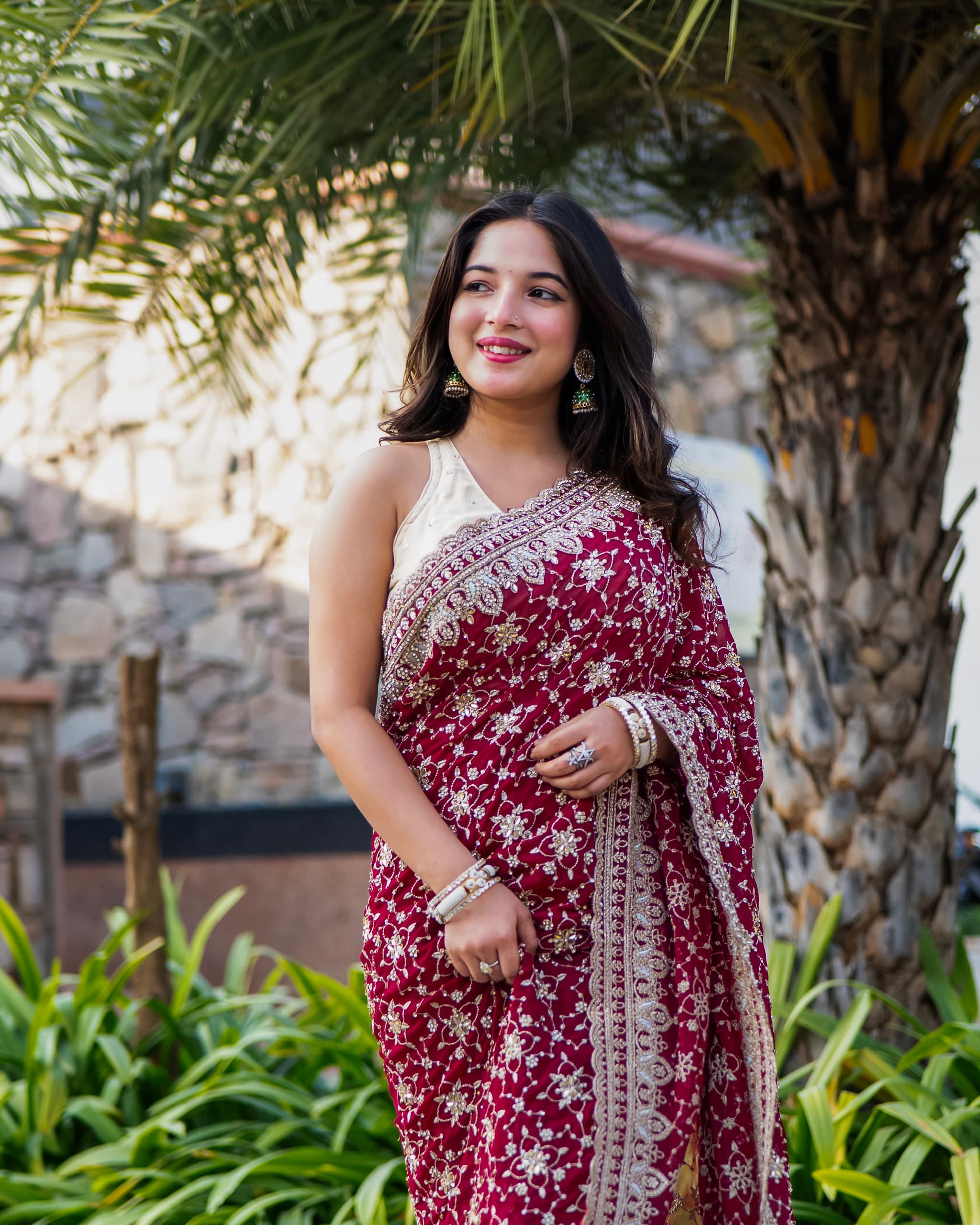 Standing beside a palm trunk in a red saree, bougainvillea and a dry stone wall filling the frame behind