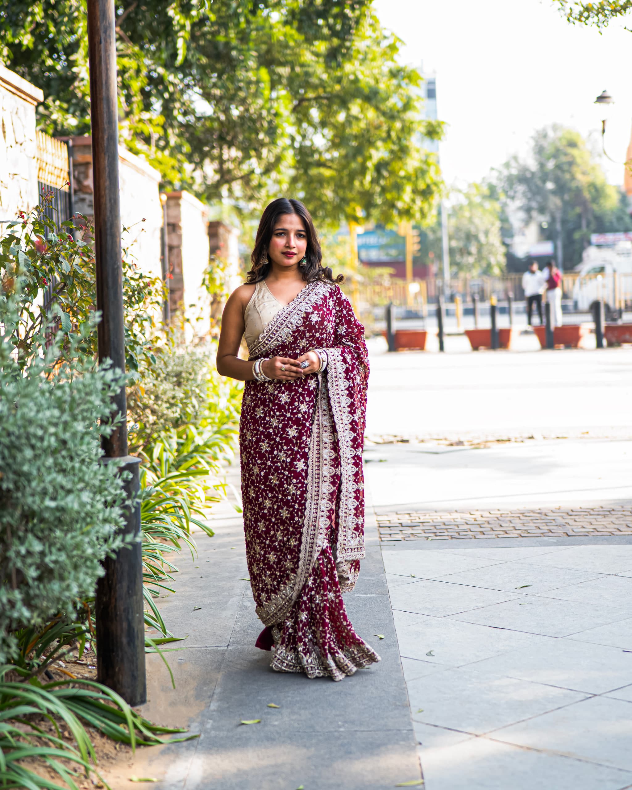 Walking away down a tree-lined street in a red saree, pavement receding into soft afternoon light