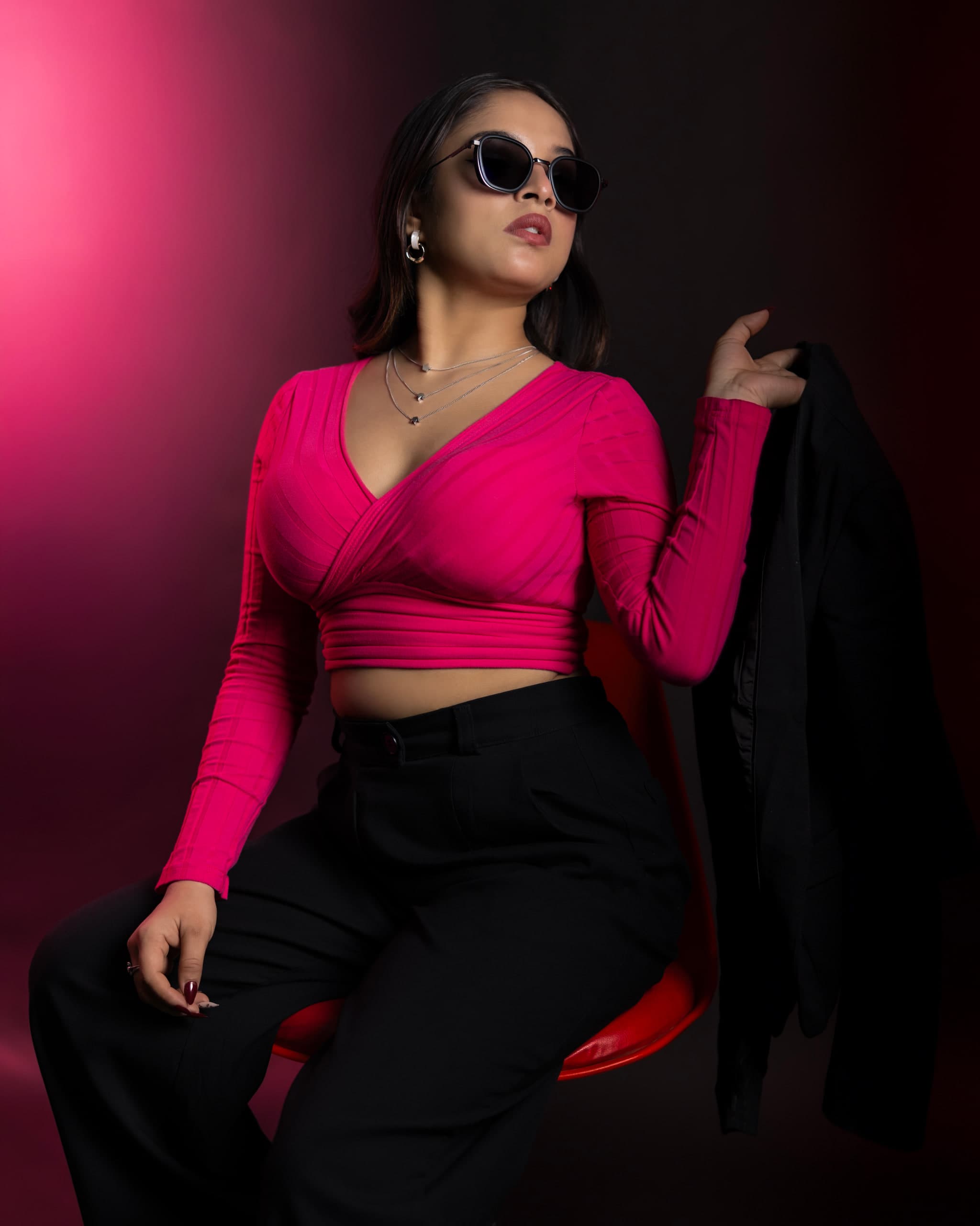 Standing on crimson in sunglasses, black blazer held off one shoulder, pink top and wide black trousers