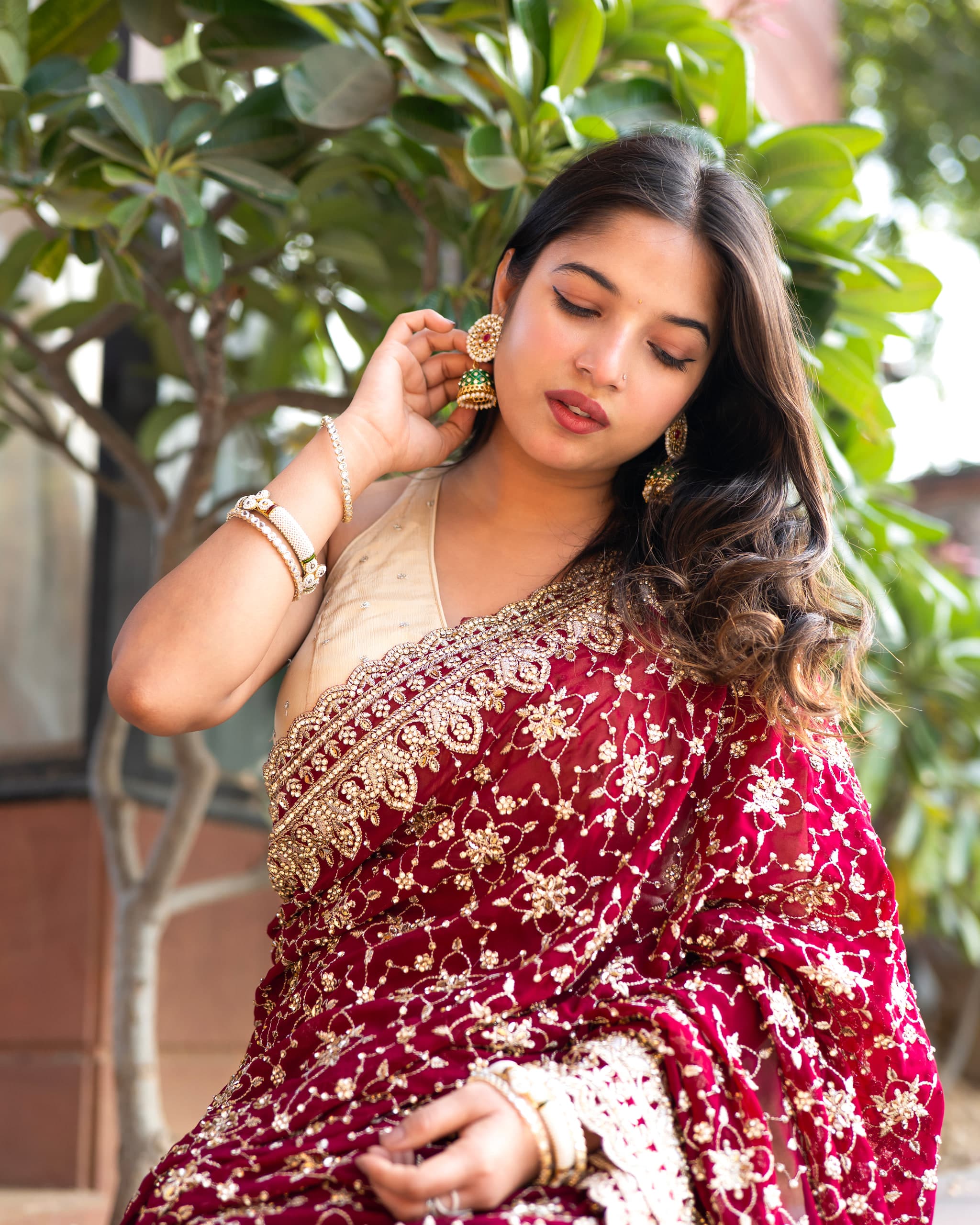 Close frame in a gold-bordered red saree, one hand raised near the face, greenery soft behind