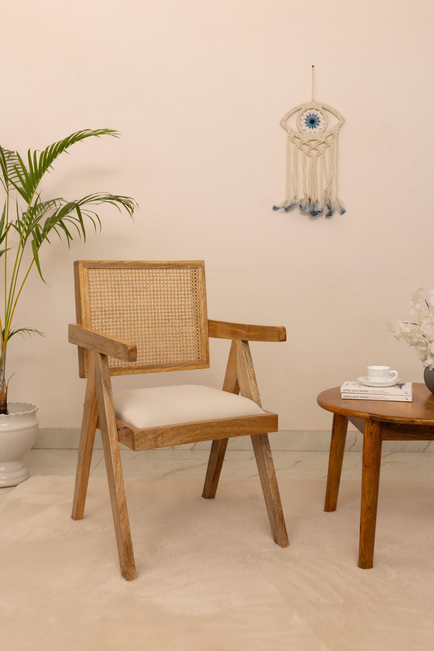 Teak and cane armchair beside a round side table, a macramé eye hanging on the pale wall