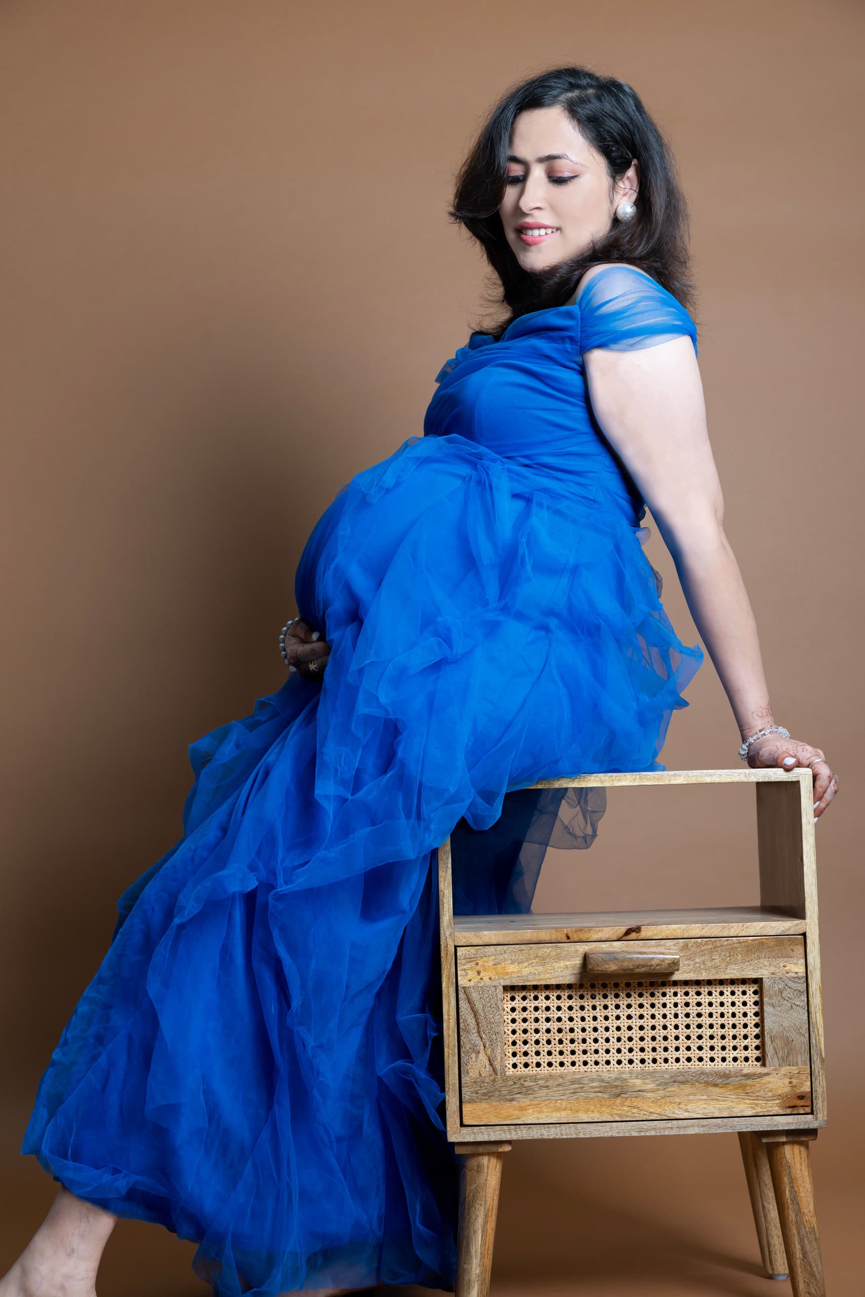 Expectant mother in a blue tulle gown against a warm brown backdrop, hand resting on her bump