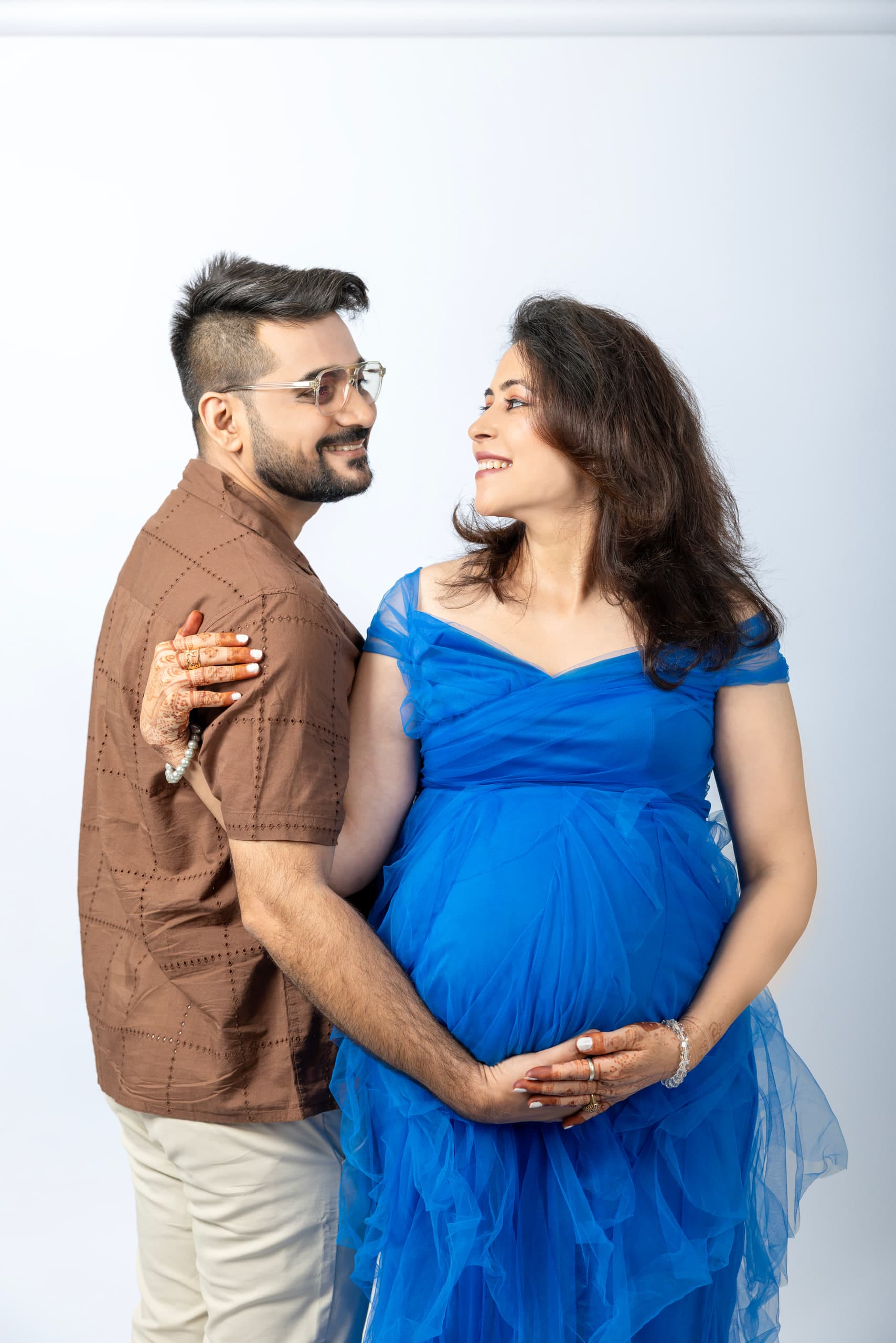 Expectant couple against white, his hand on her shoulder, both their hands on the bump