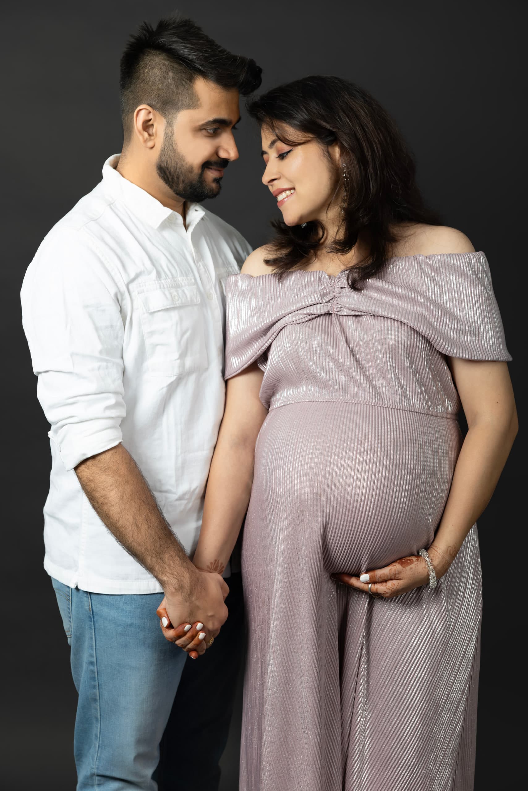 Expectant couple against charcoal, holding hands, her other hand cupped under the bump