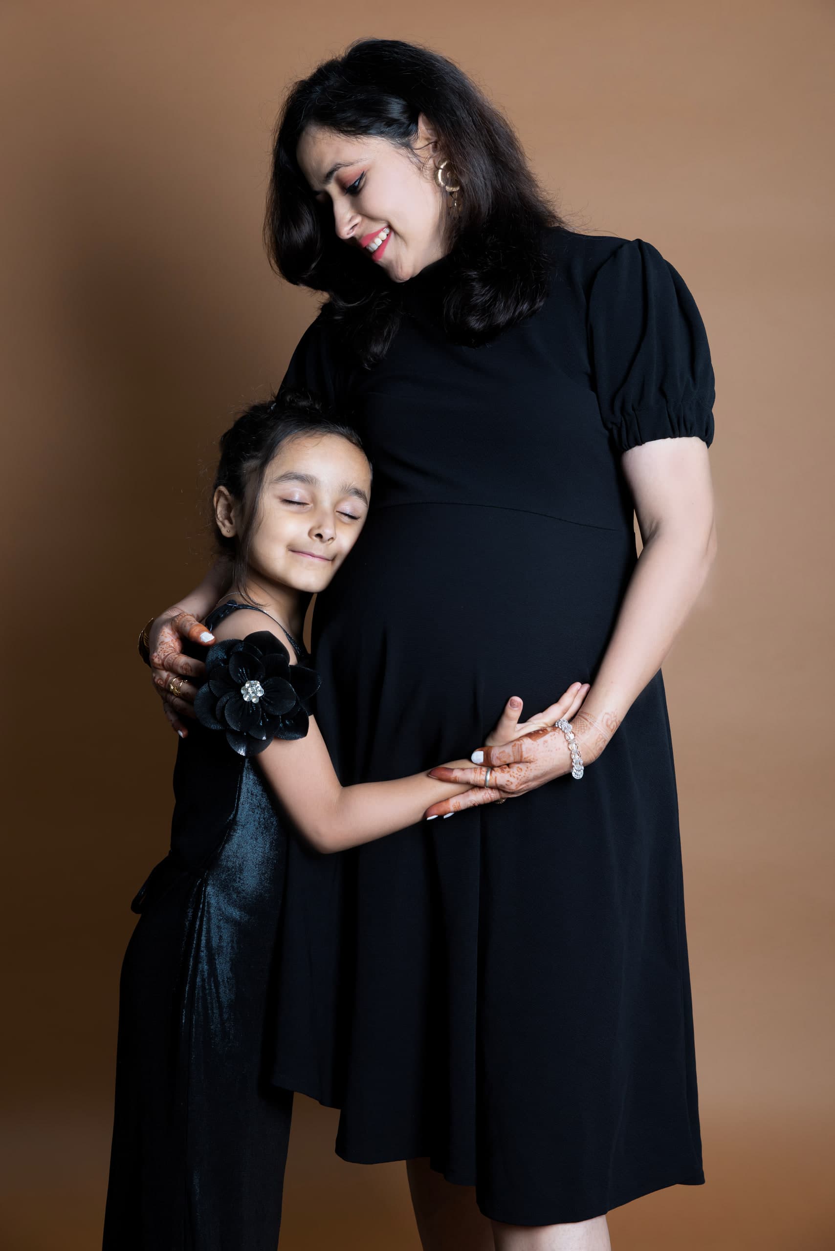 A girl leans into her expectant mother, both in black, their hands together over the bump