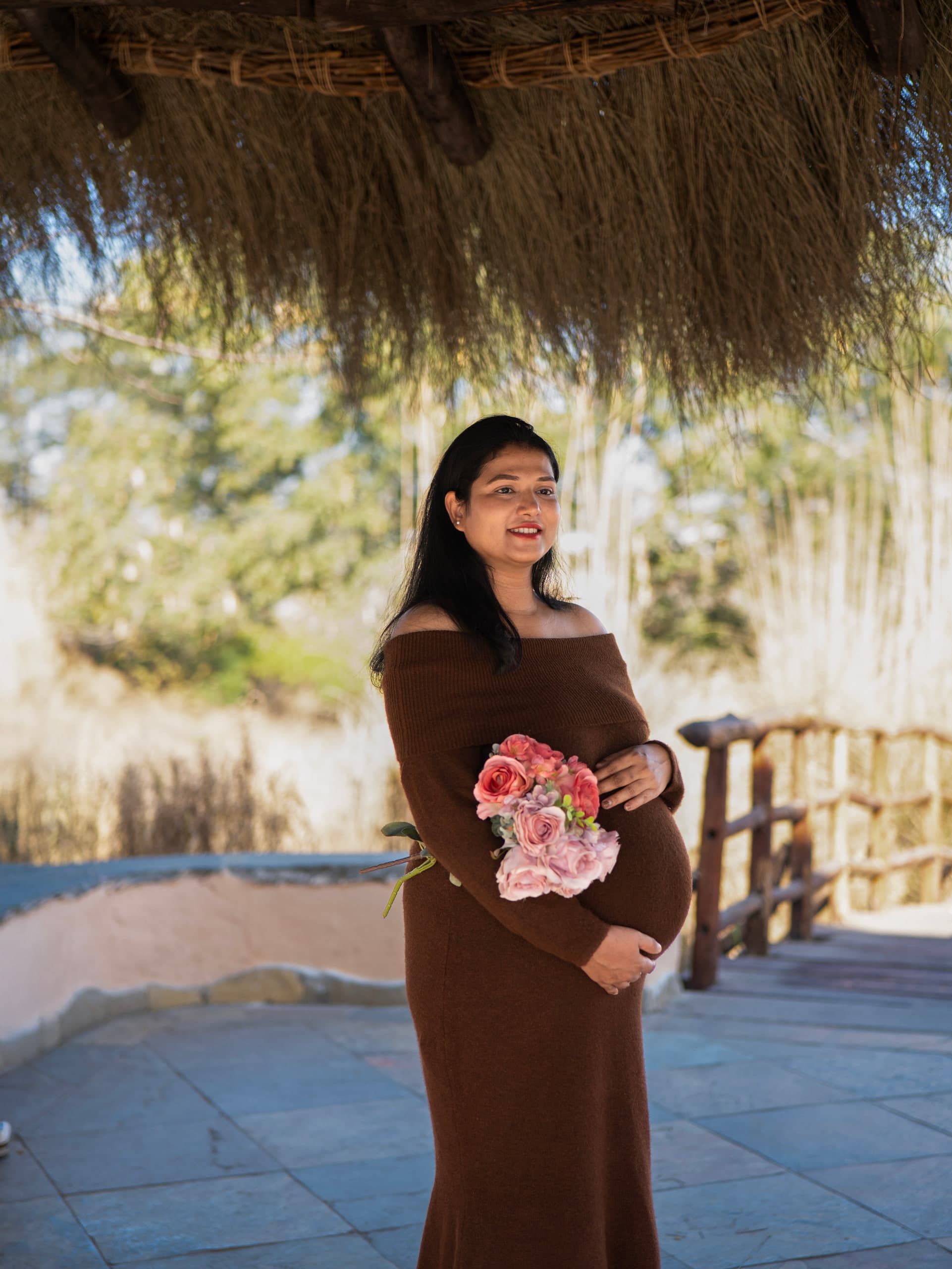 Expectant mother outdoors in brown holding a posy, tall reeds behind her