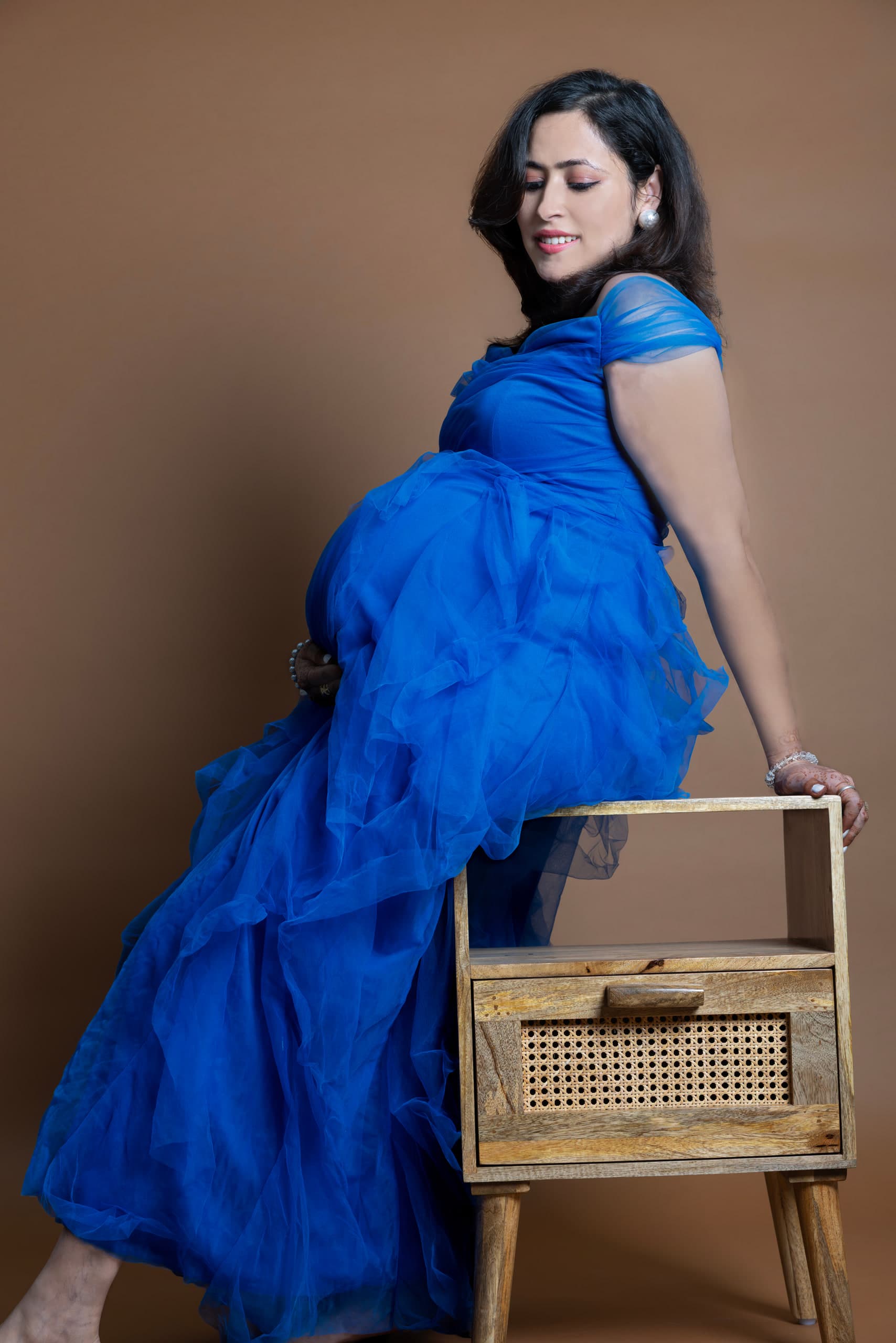 Expectant mother in blue tulle leaning on a cane chair against a brown ground
