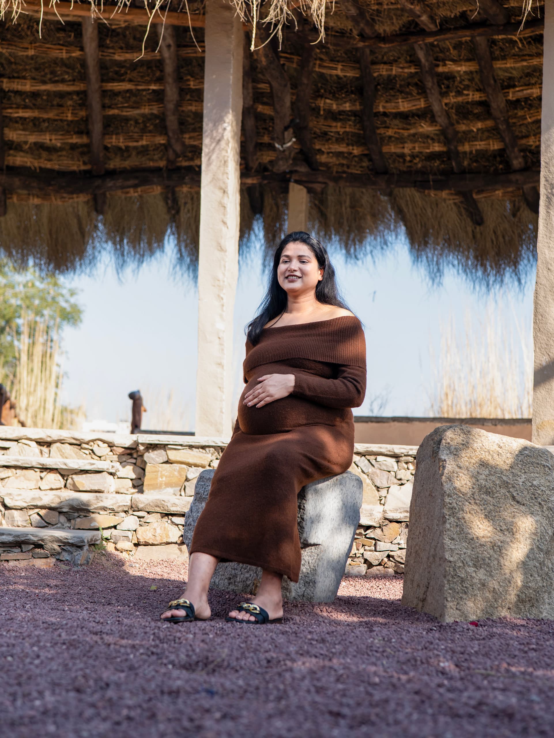Expectant mother seated under a thatched shelter, dry stone wall behind