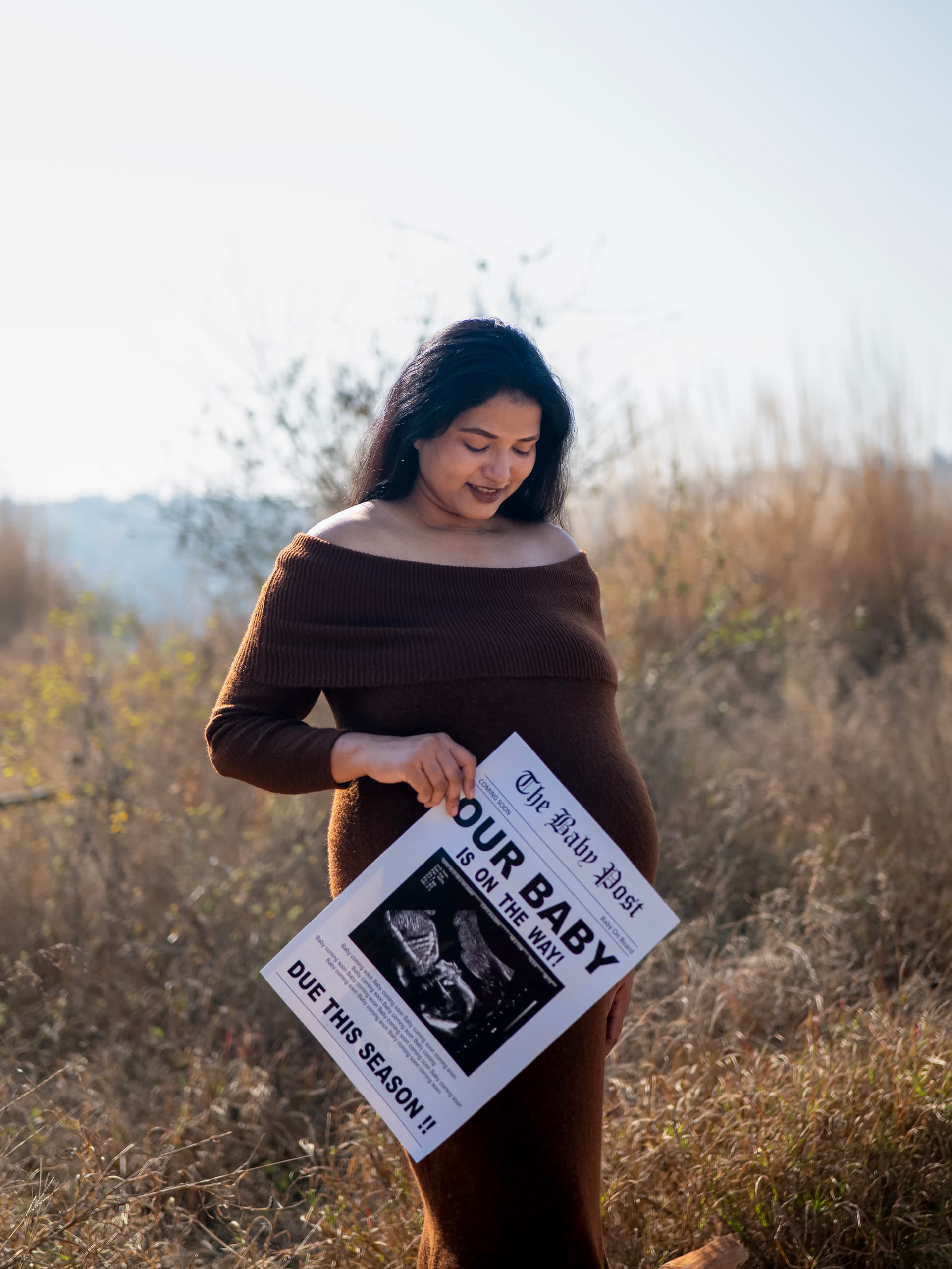 Expectant mother in a field holding a hand-lettered board reading "our baby"