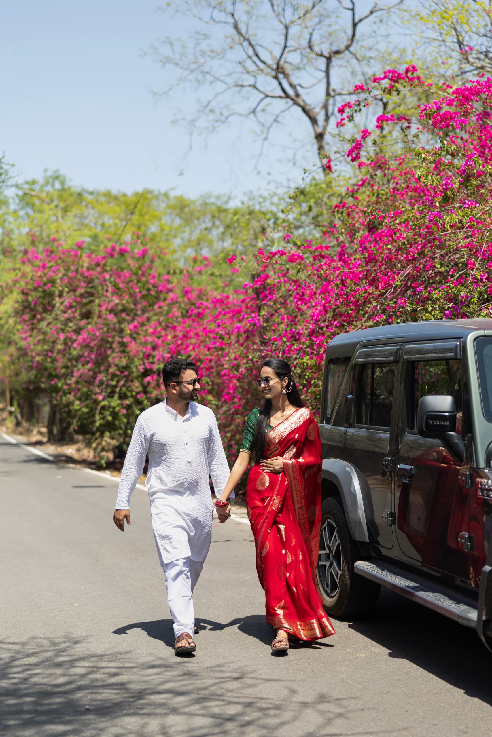 Couple walking hand in hand along a road beneath flowering bougainvillea, the car behind them