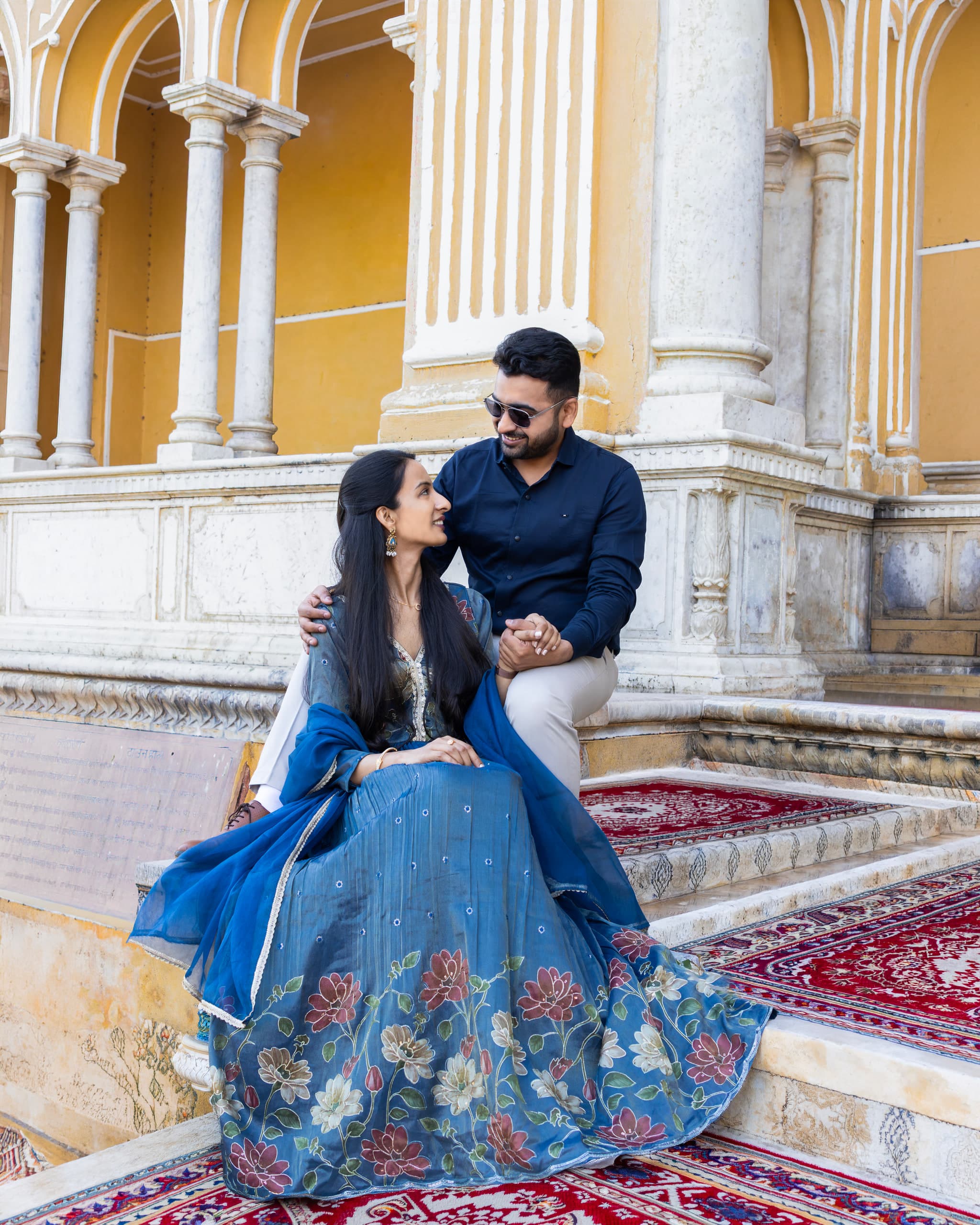 Couple seated on the steps of a colonnaded palace, blue and gold lehenga spread across a red carpet