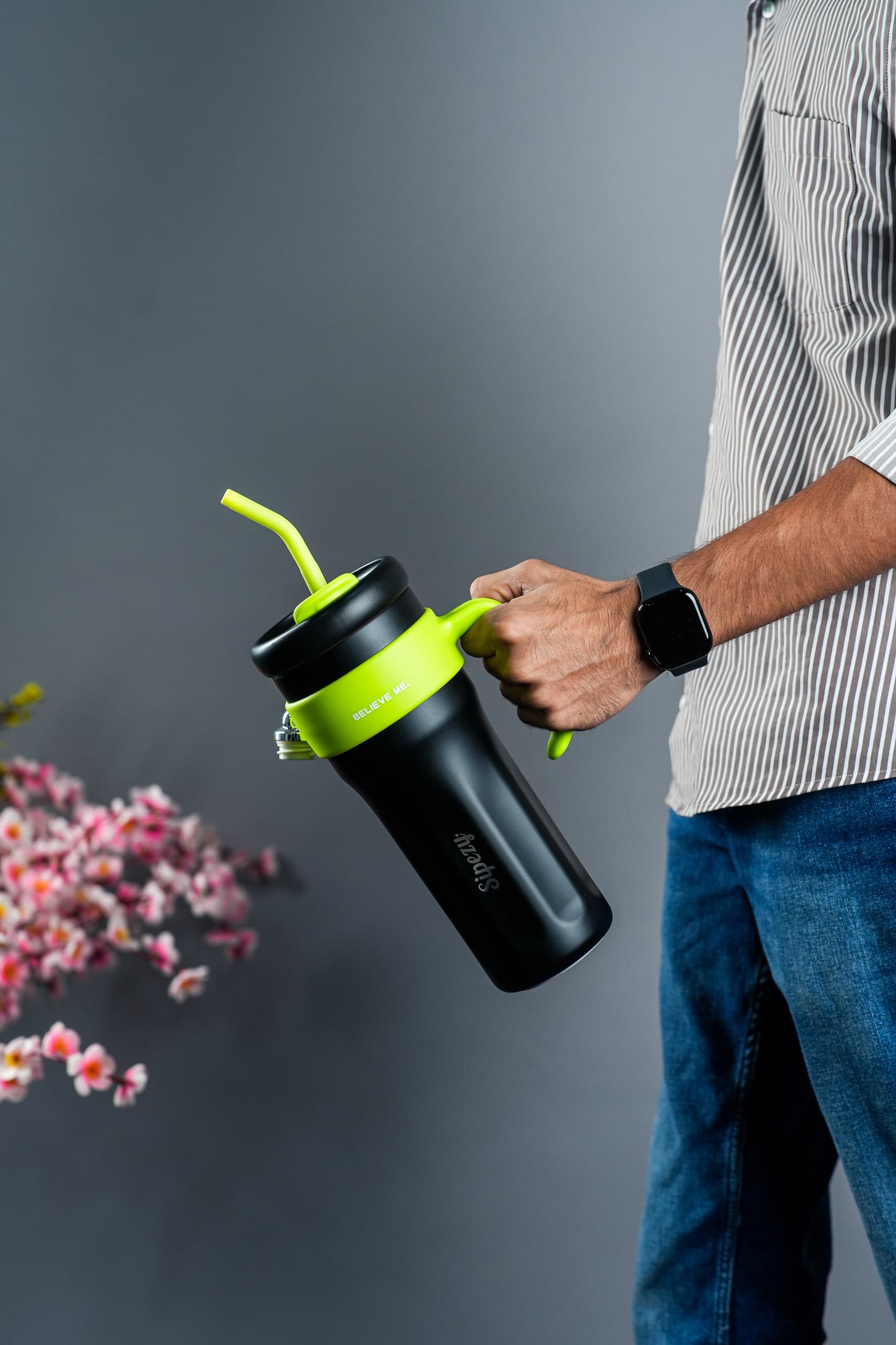 Hand lifting a black insulated tumbler by its handle, blossom soft behind on grey