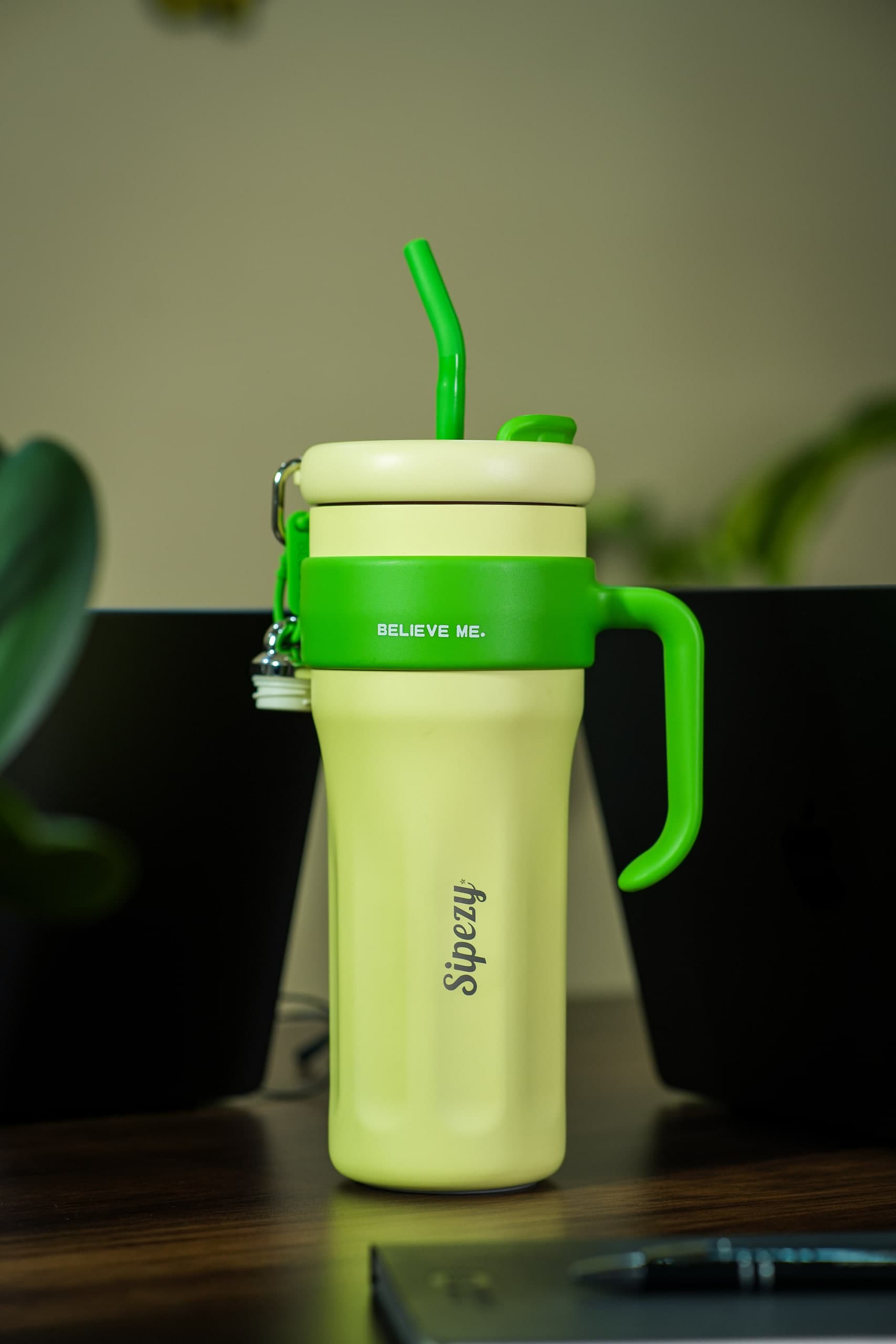 Lime-green tumbler on a desk beside a pen, plants behind