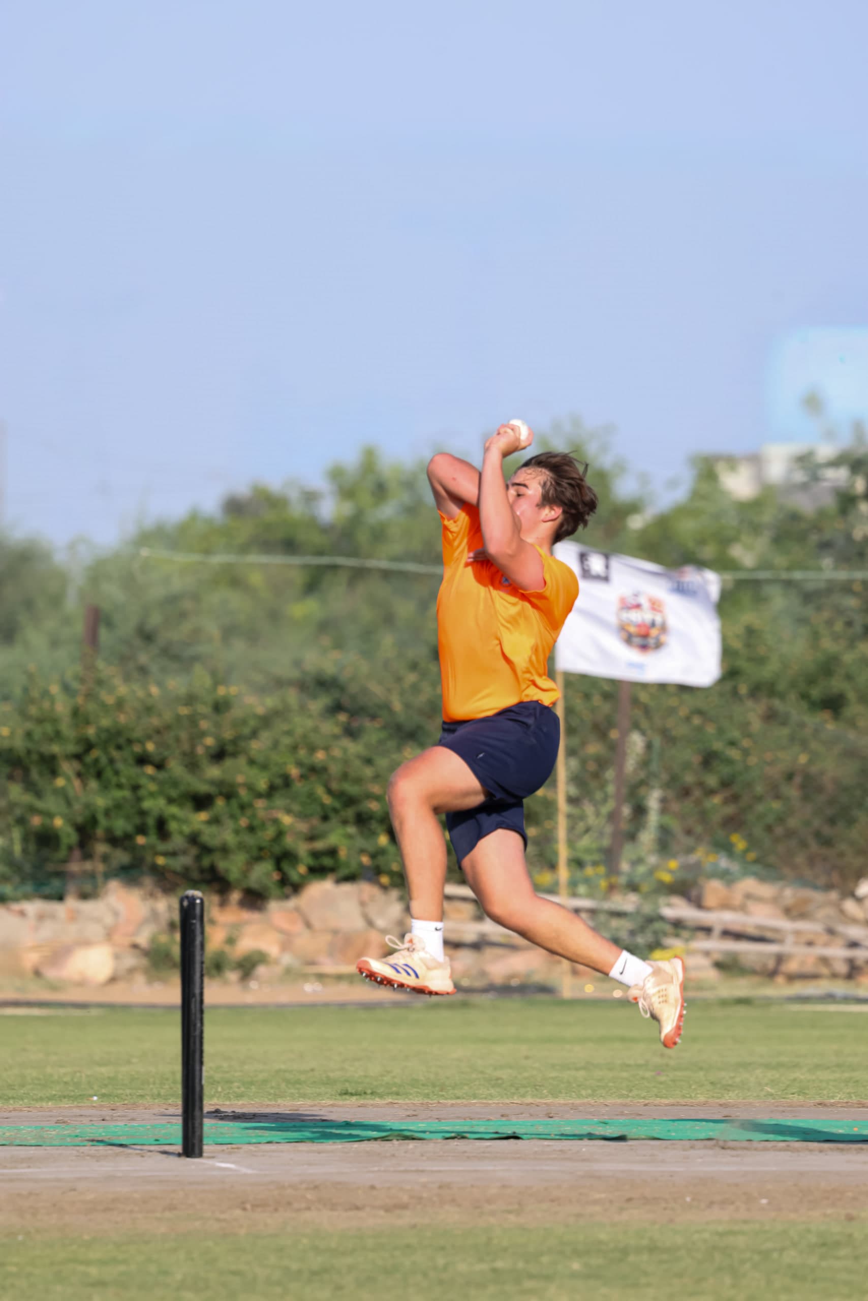 Bowler at the top of his leap against a bright sky, front arm high, boundary boards behind