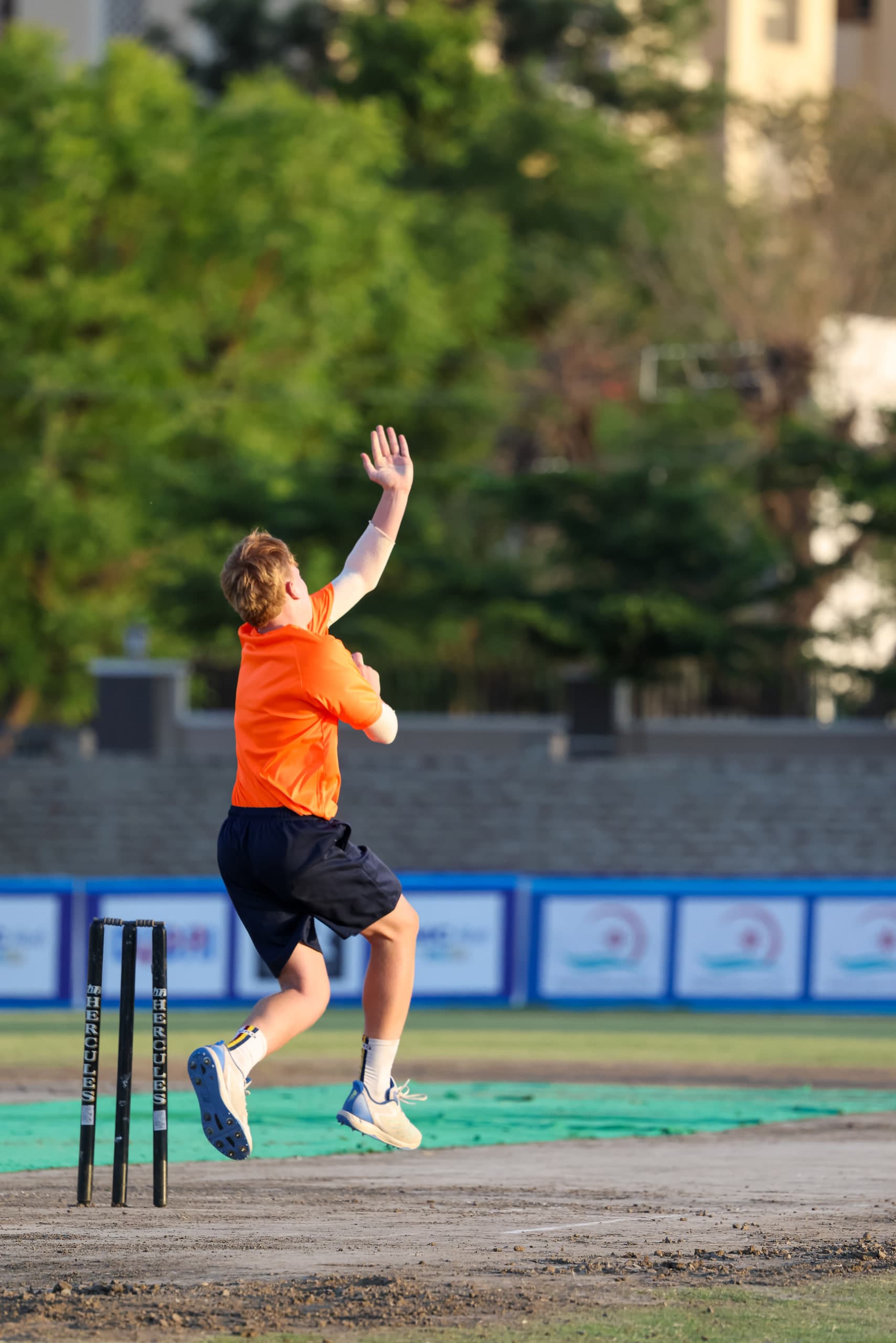 Bowler airborne in the delivery stride over the stumps, dust rising off the worn pitch