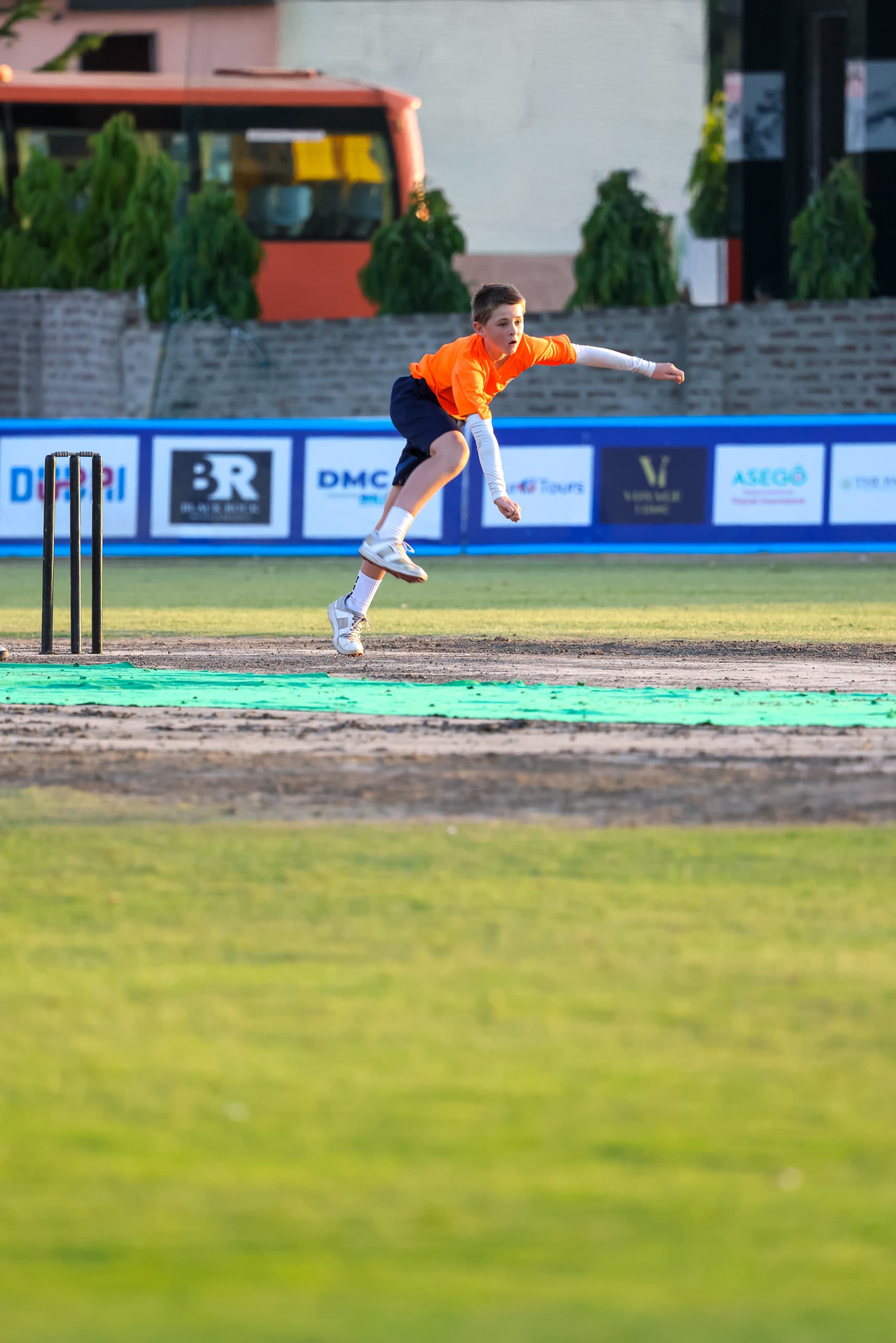 Bowler in full delivery stride across a green outfield, stumps and matting pitch ahead of him