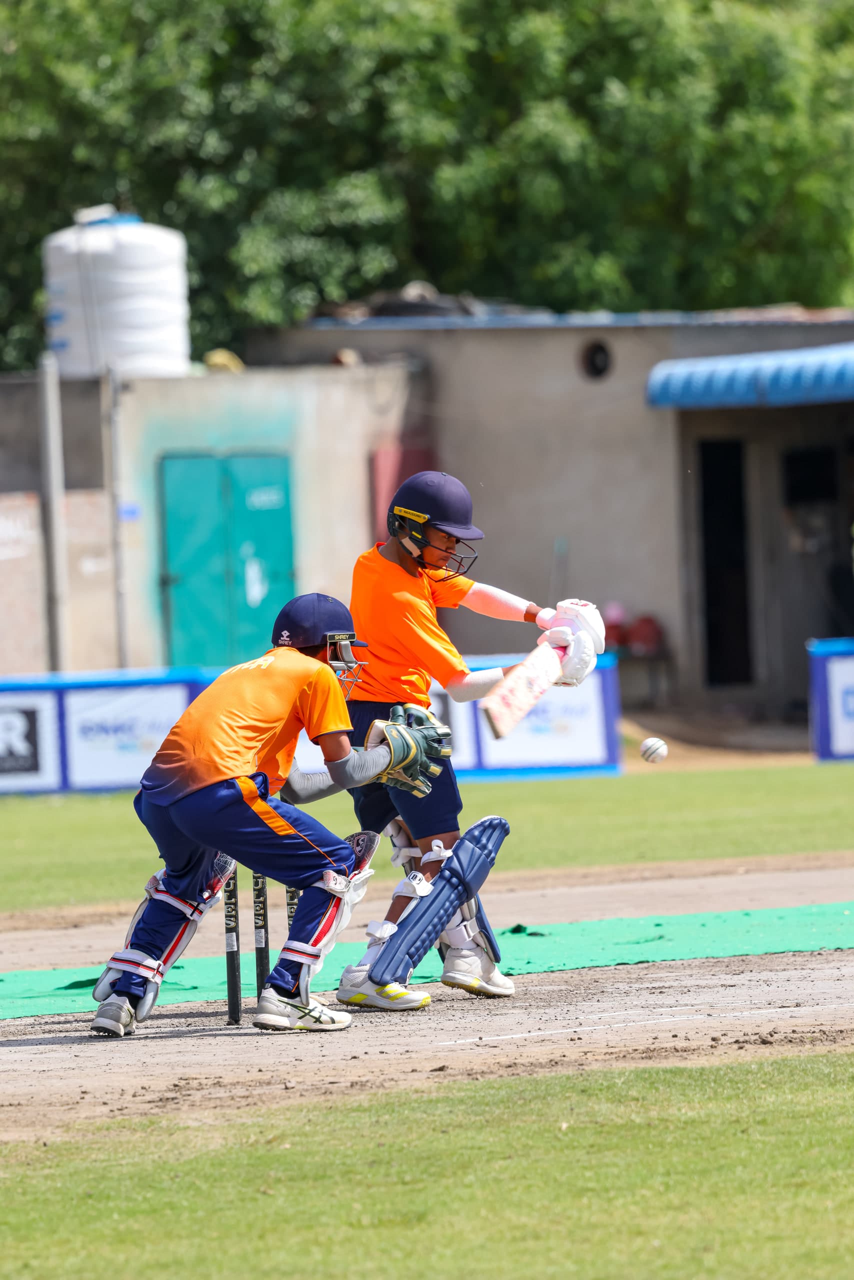 Batter driving through the line in whites, wicketkeeper in orange crouched at the stumps