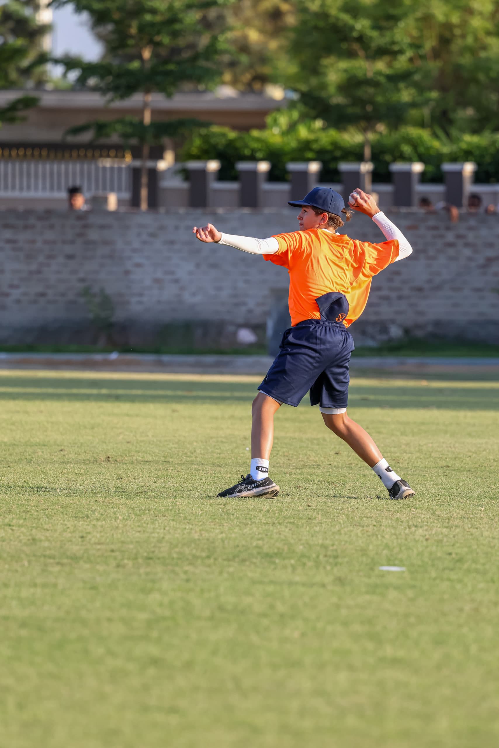 Fielder winding up to throw from the deep, arm cocked, trees and pavilion behind