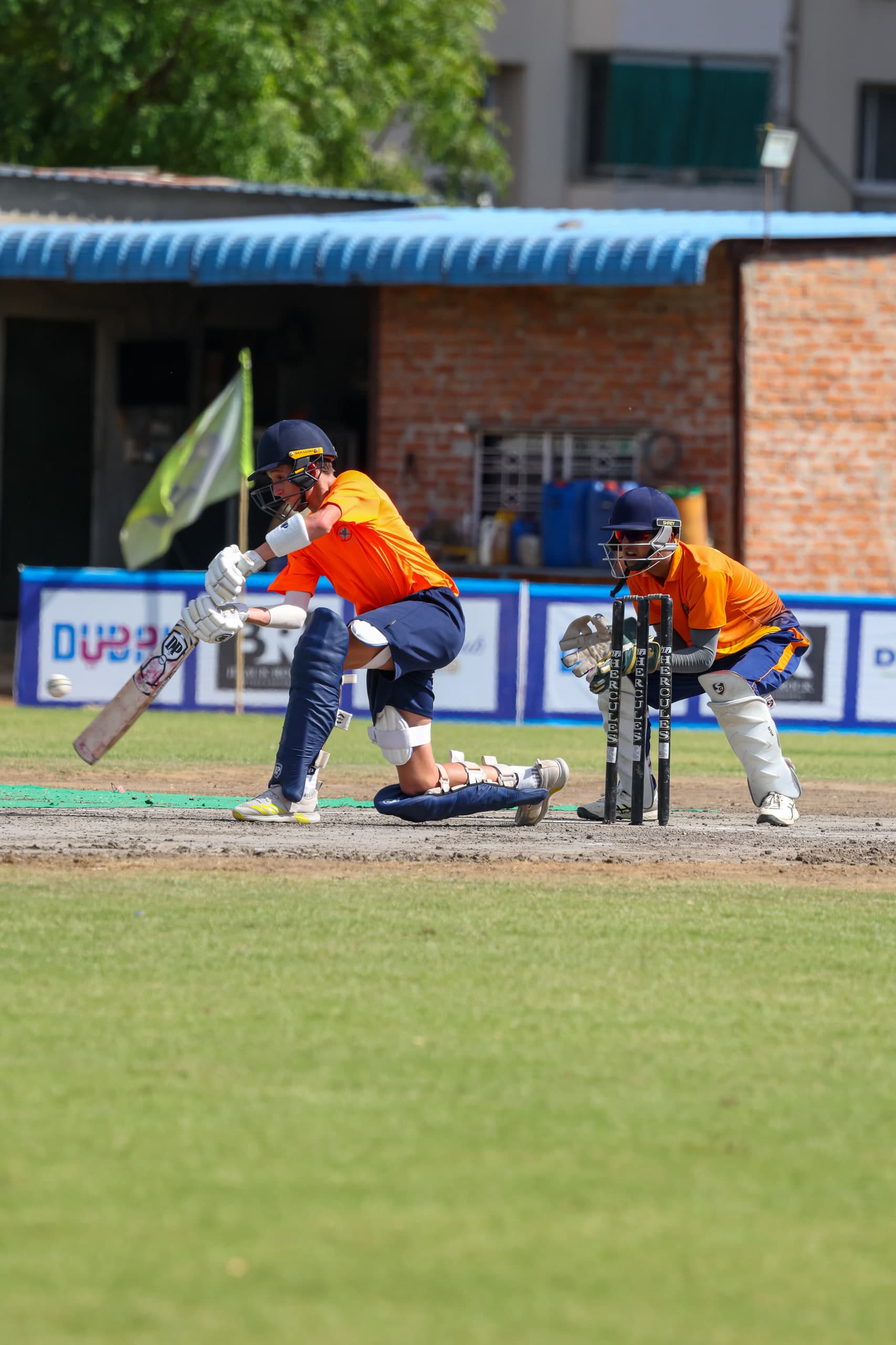 Batter, wicketkeeper and slip fielder set and waiting, brick pavilion across the ground