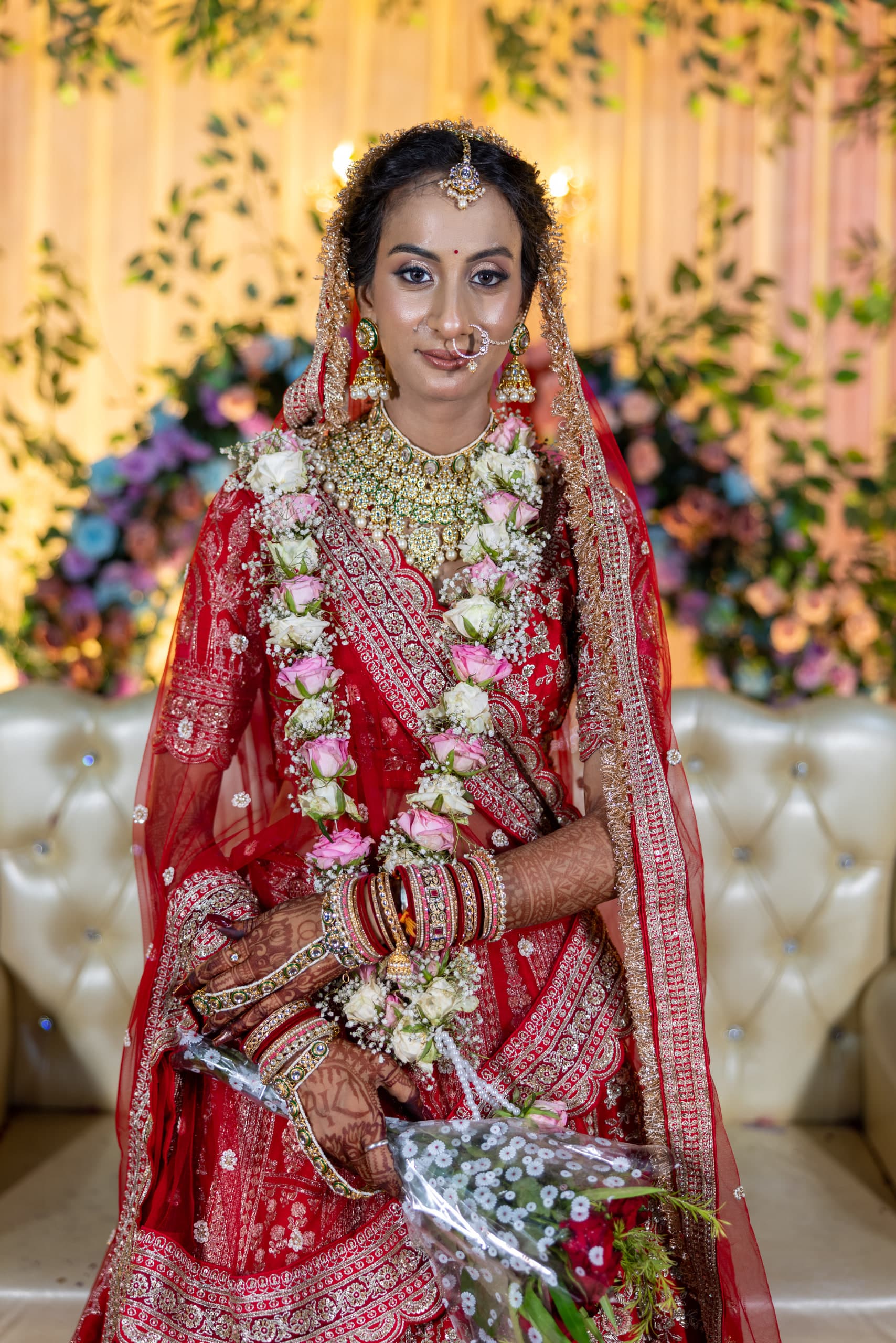 Bride standing in red, kundan necklace and nose ring, a bouquet in her mehndi-covered hands