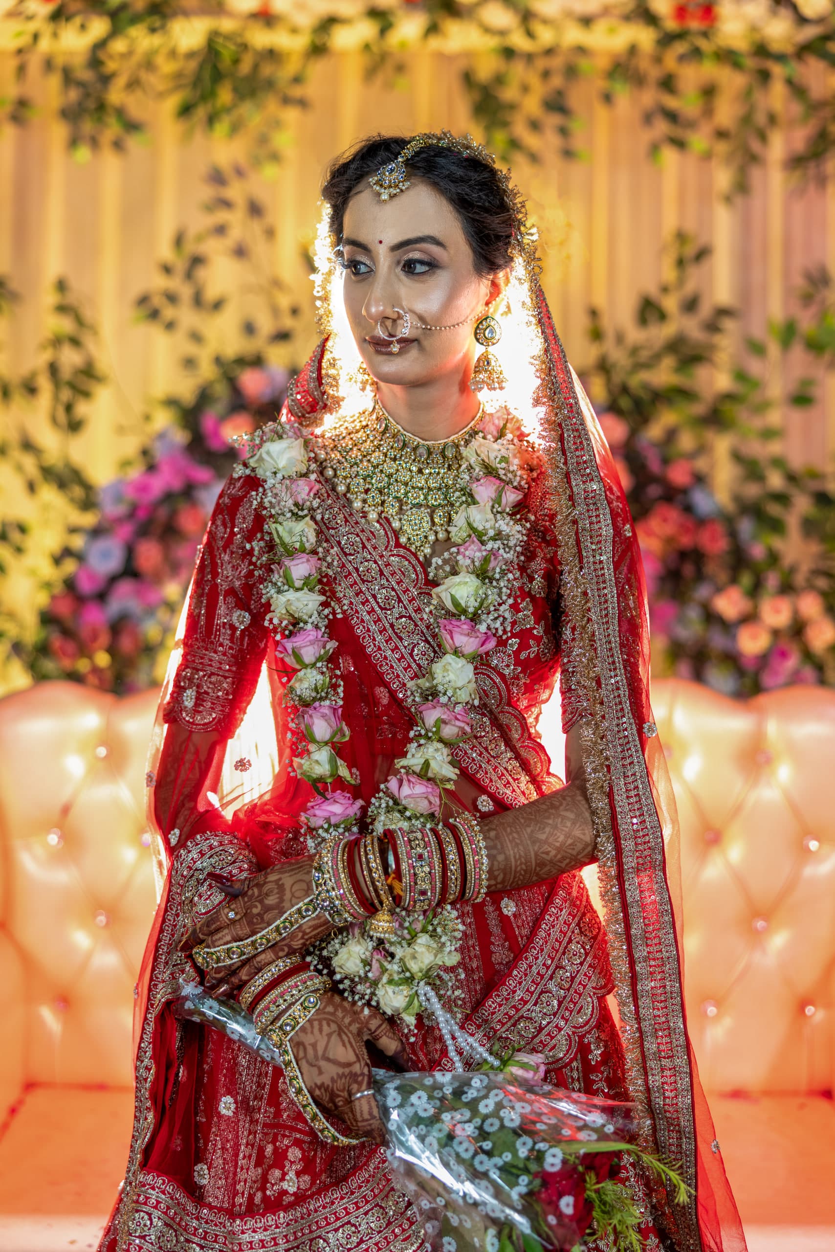 Backlit bride in red and gold, hands folded over a white bouquet, flowers and leaves behind her