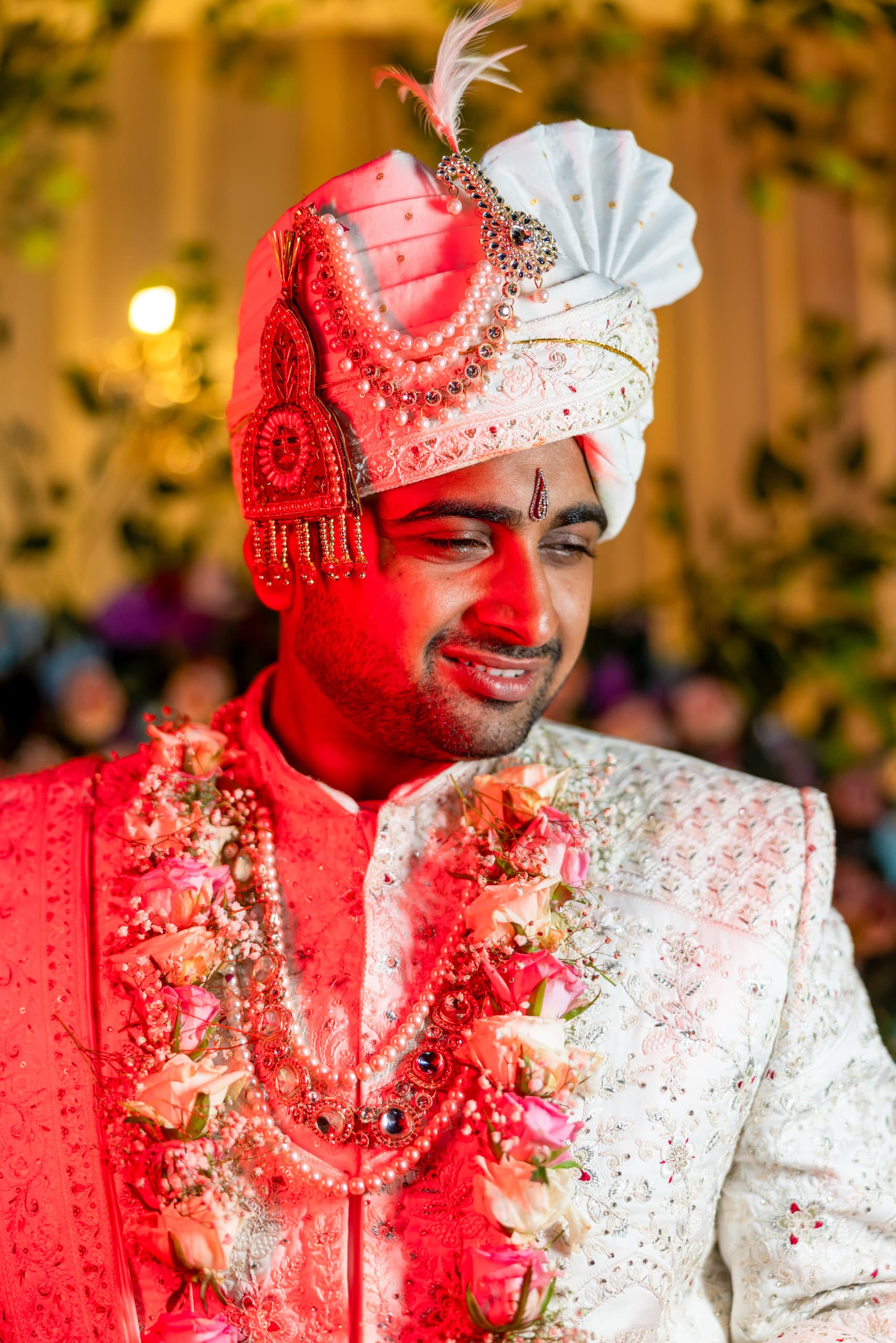 Groom smiling under a red and ivory safa, the whole frame washed red by the light through the tent