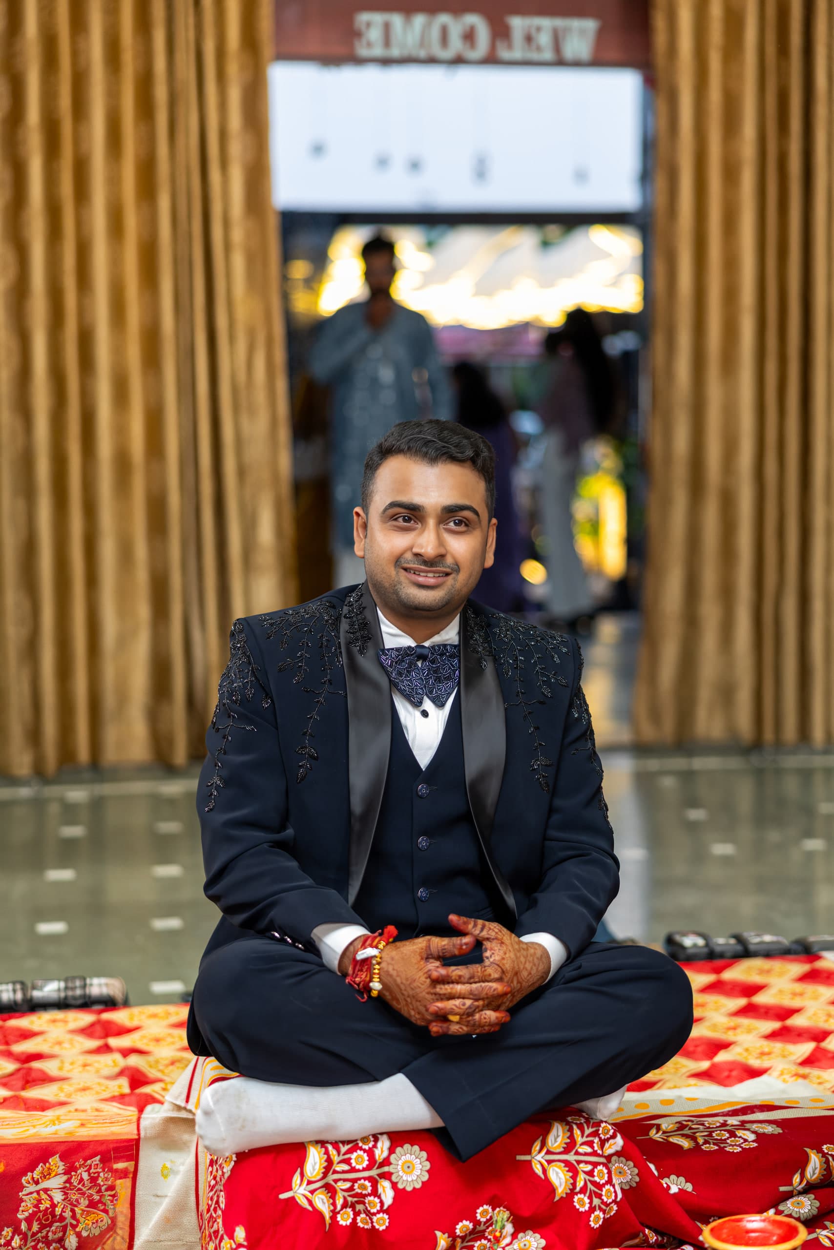 Groom cross-legged on a red floor spread in a navy tuxedo, the venue doorway open behind him