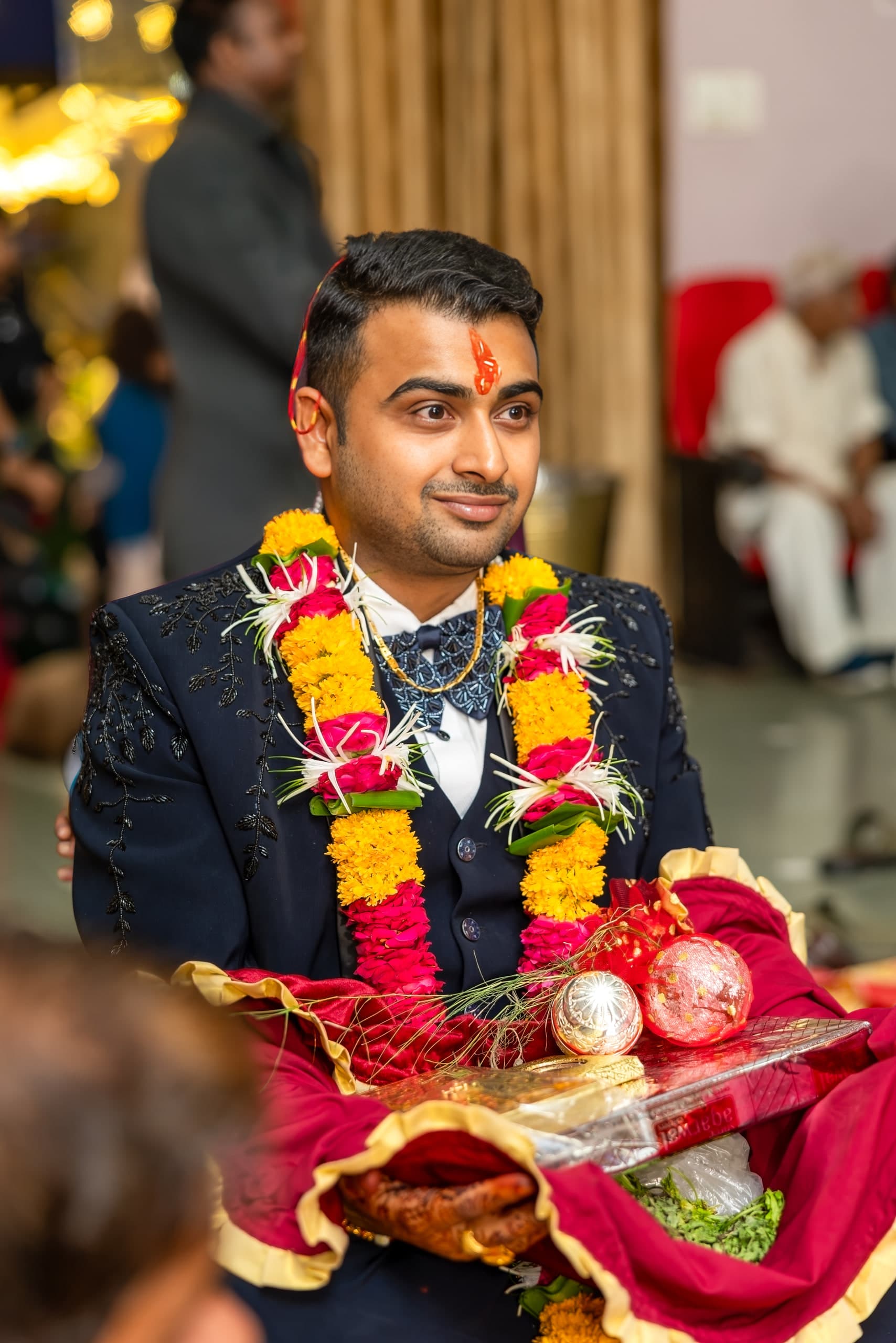 Groom carrying a covered ritual thali, marigold garland over a navy jacket, guests seated behind