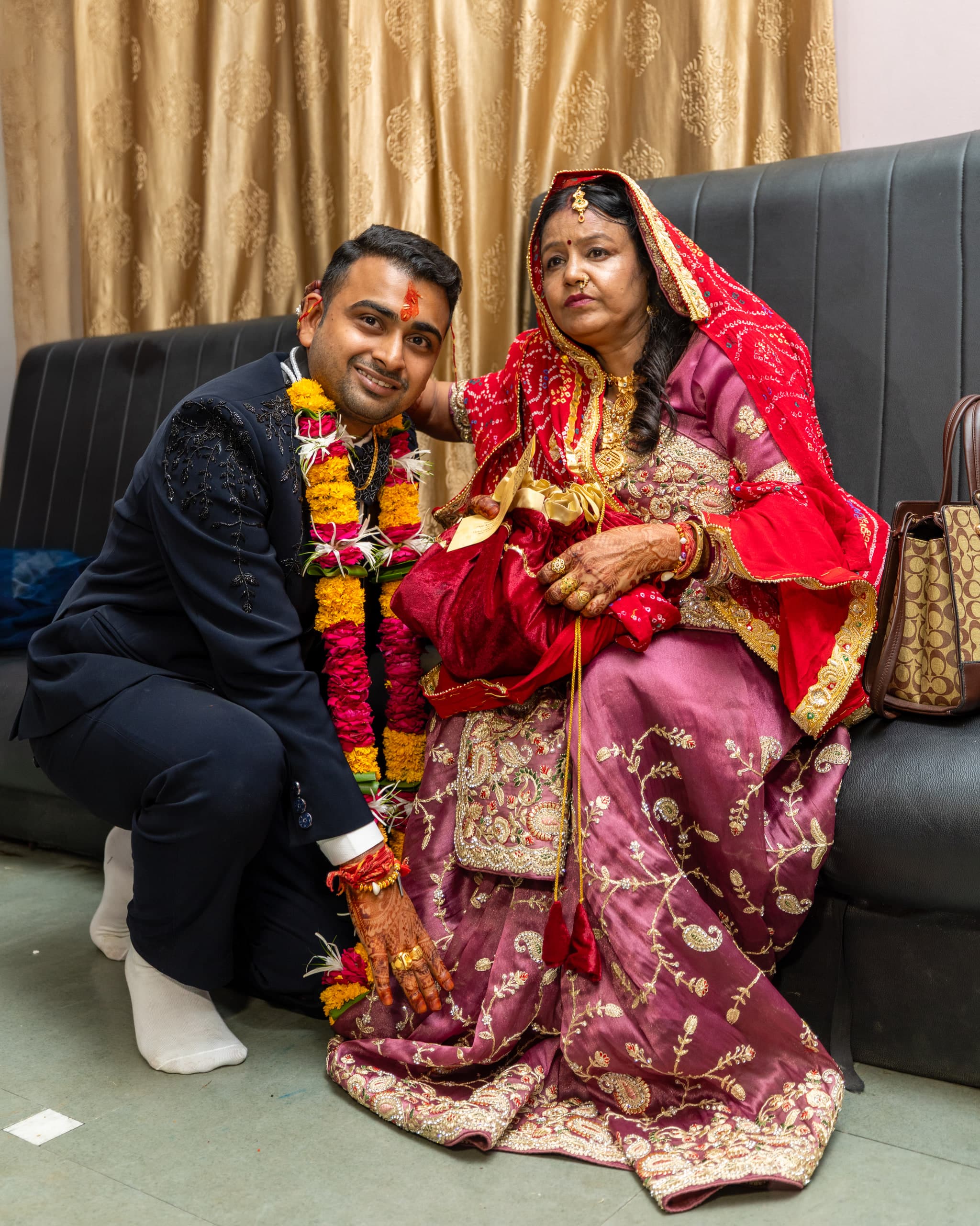 Groom crouches beside an older relative seated in a magenta sari, both turned to the camera