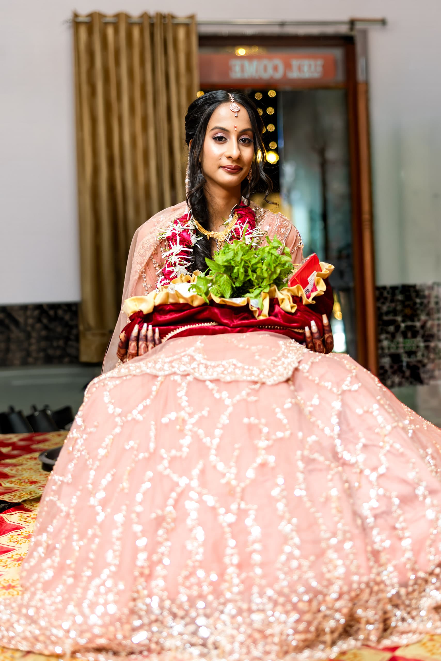 Woman in a blush lehenga seated behind a tray of ritual greens, mehndi dark on both hands