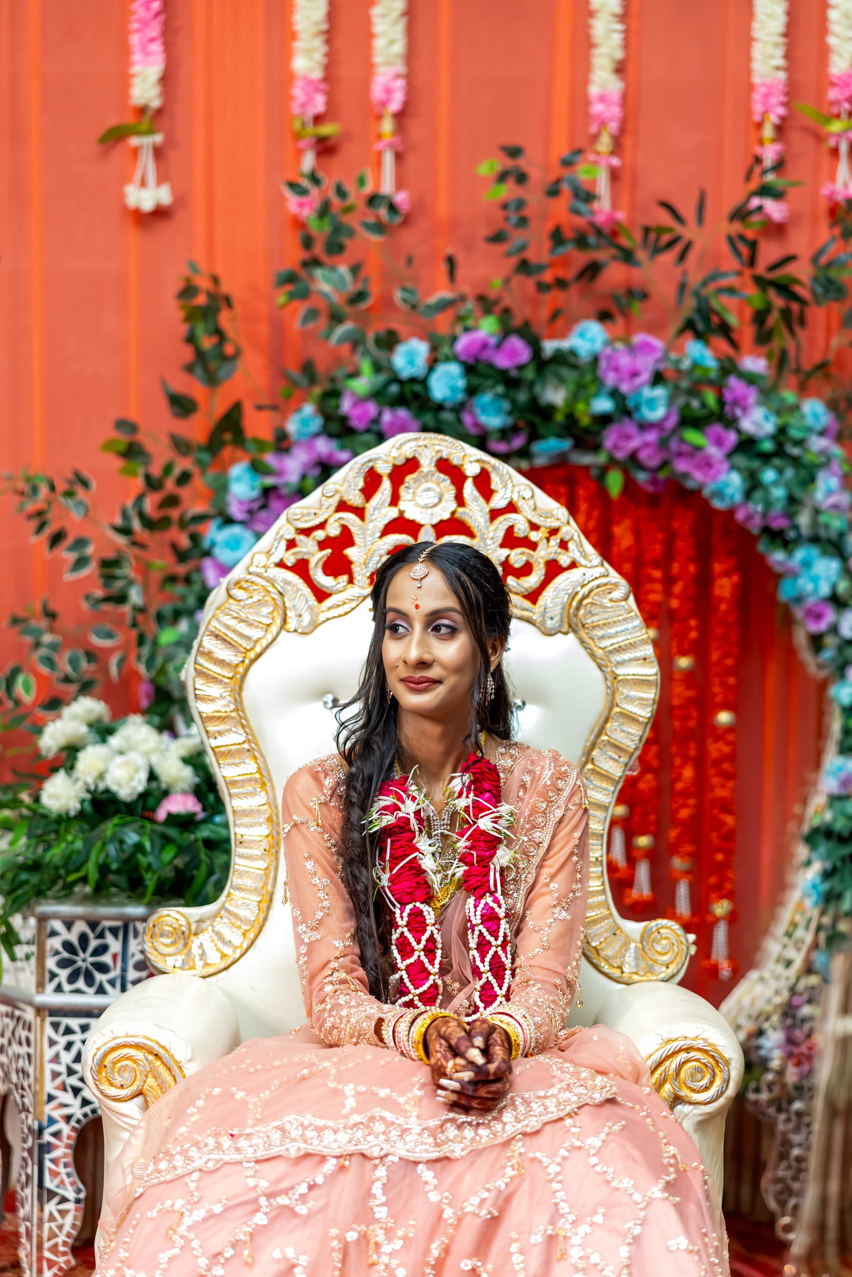 Woman in blush and gold on a carved white and gold chair, drapes and flowers behind her
