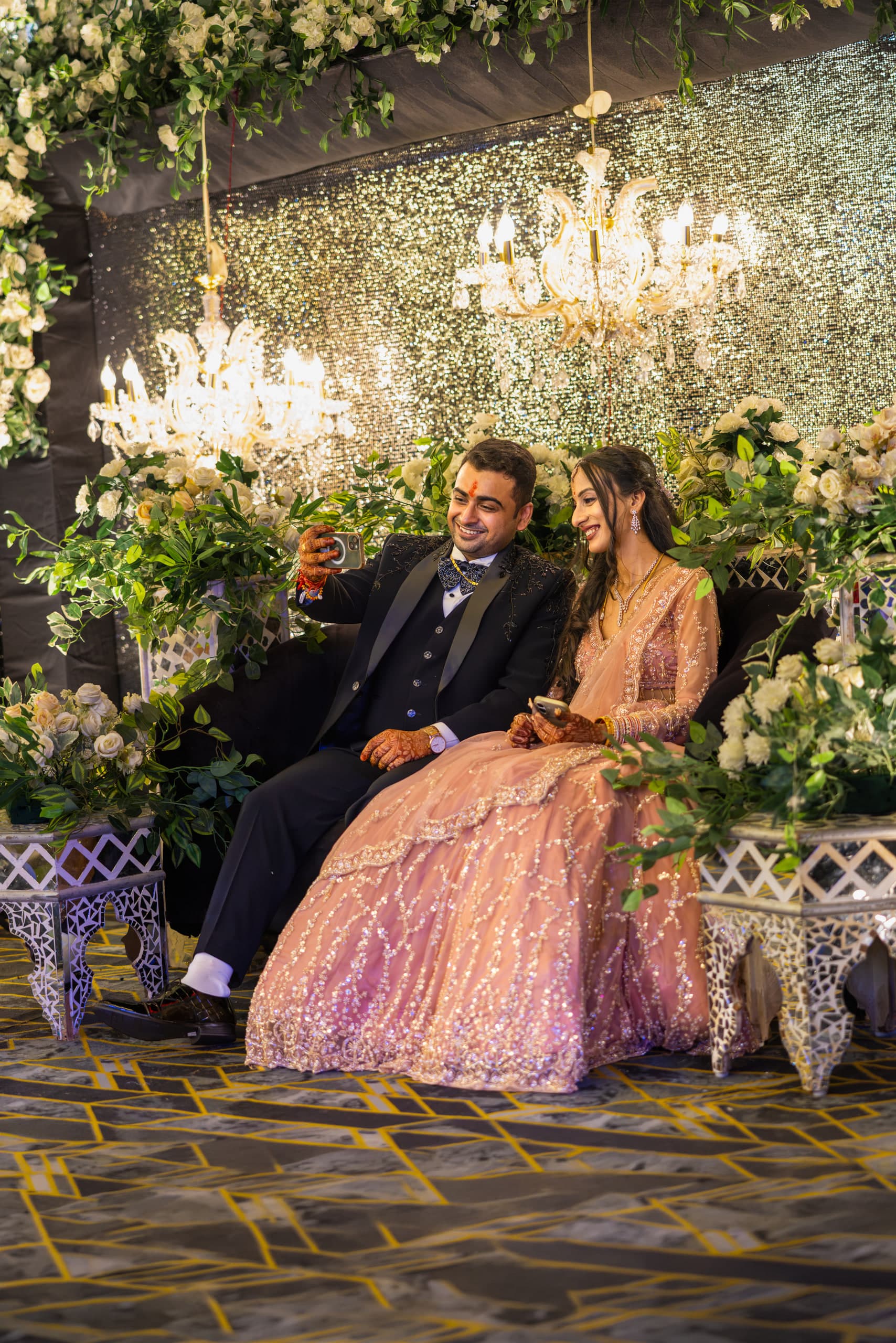 Couple on a low sofa under chandeliers and a wall of white flowers, both of them laughing