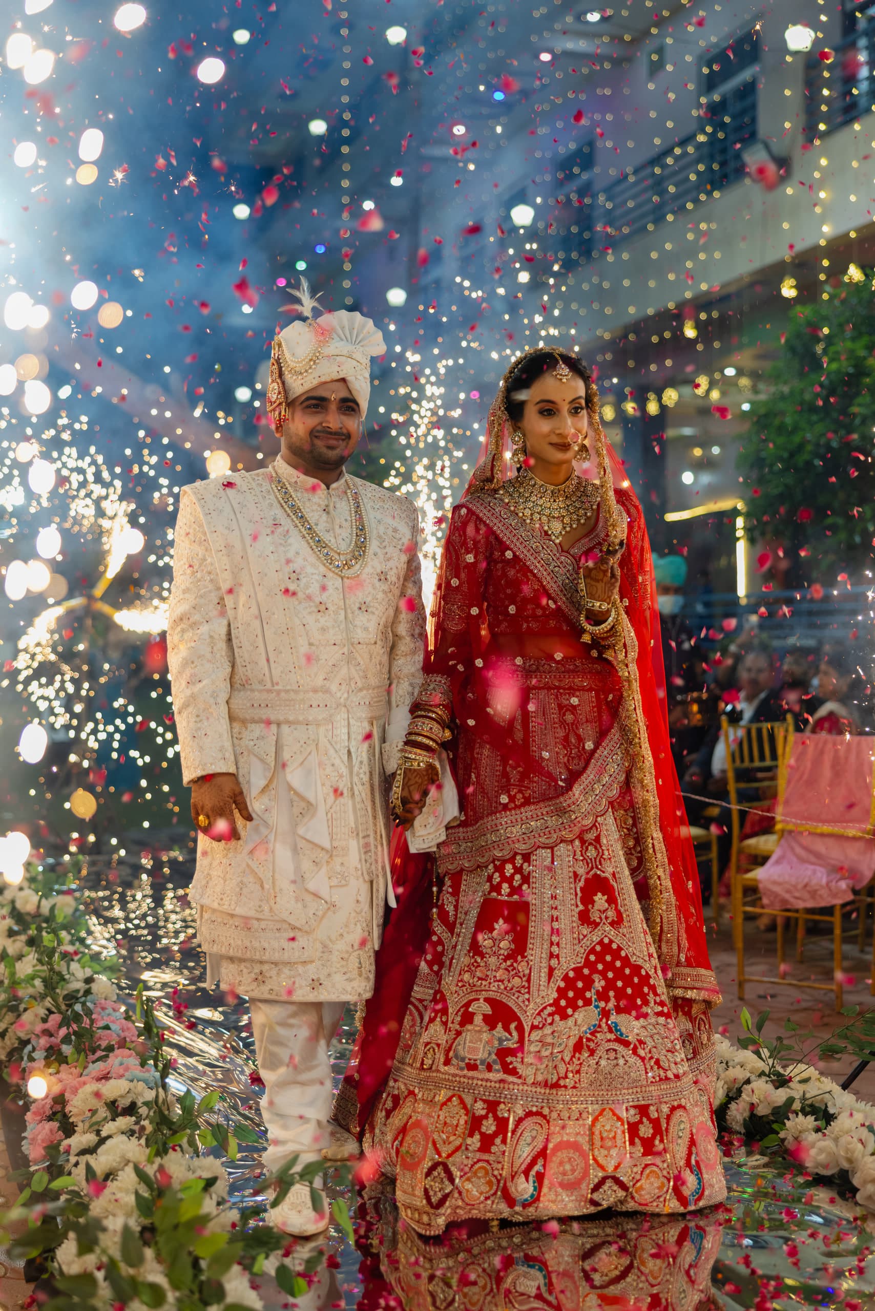 Couple walking through falling confetti, he in an ivory sherwani, she in red