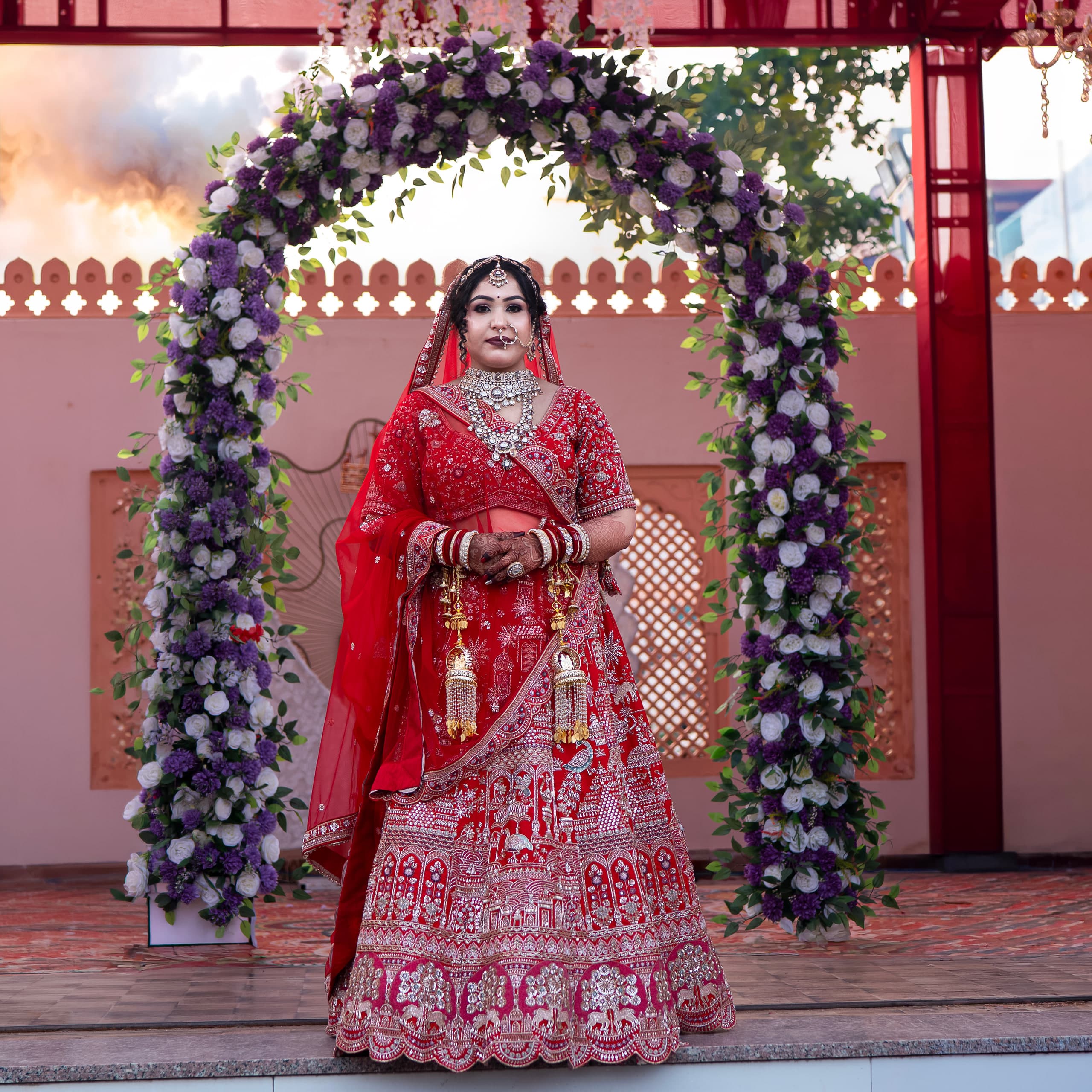 Bride beneath a purple and white floral arch, a pink palace facade behind her