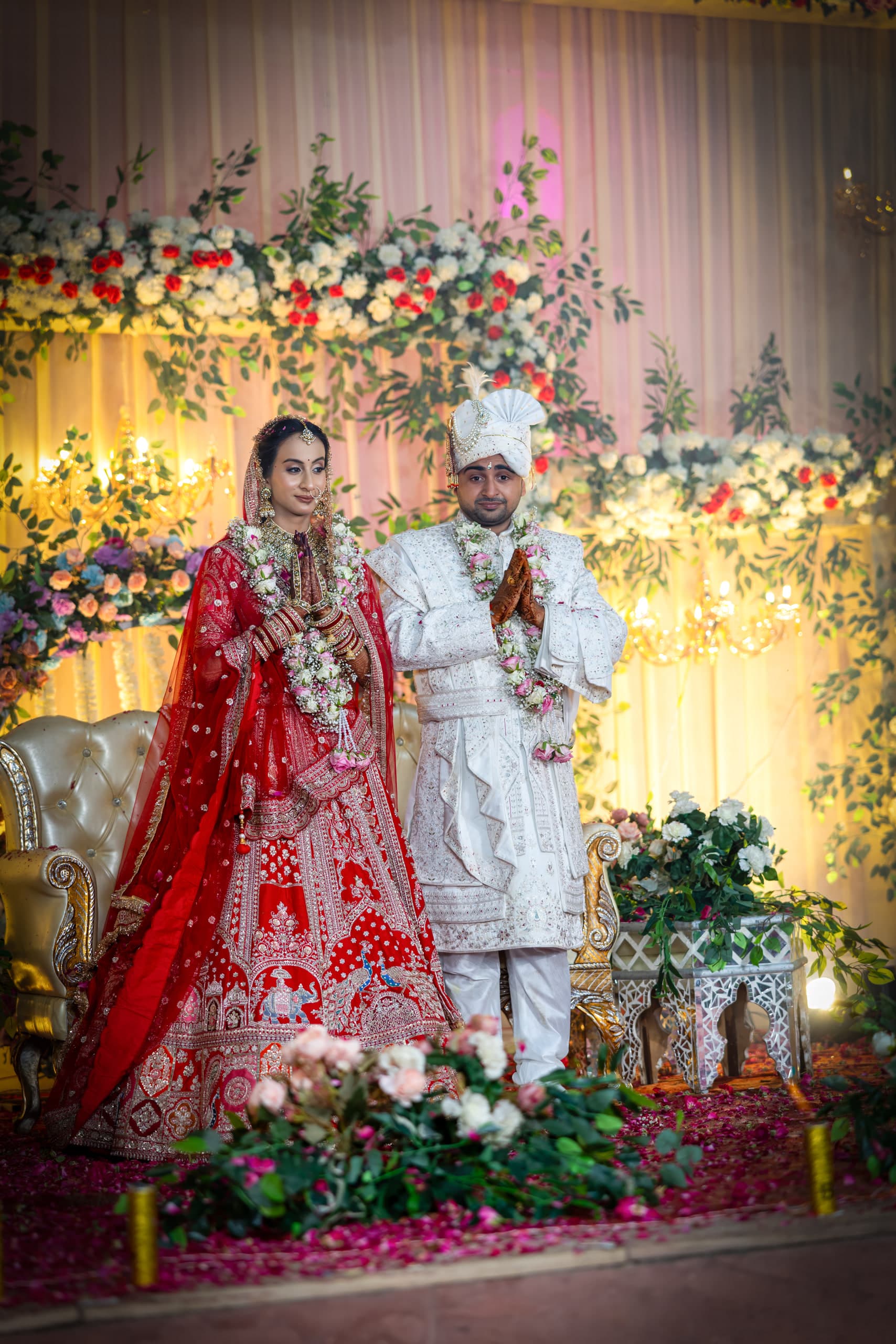Couple on a flower-banked stage, red lehenga beside an ivory sherwani